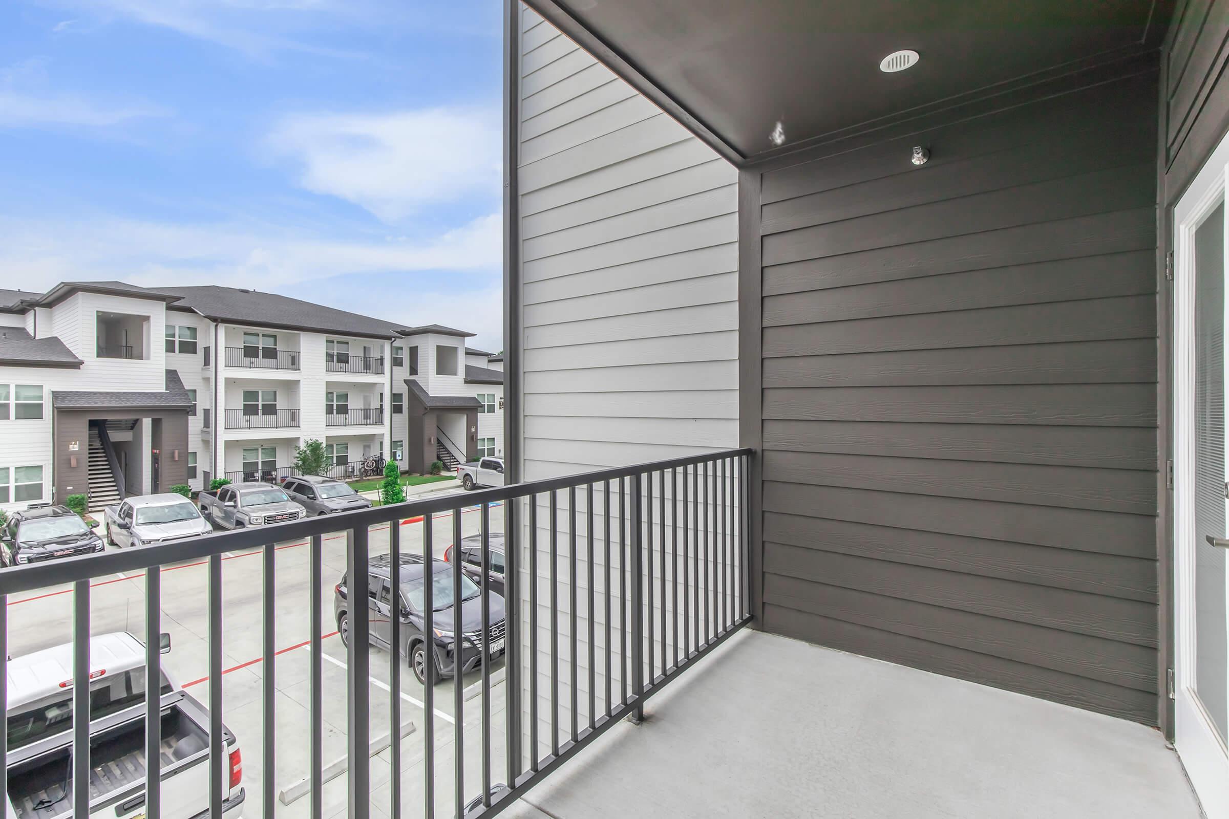 A view from a balcony featuring a railing, overlooking a parking area with parked vehicles and modern apartment buildings in the background. The sky is partly cloudy, and the architecture showcases a contemporary design with a mix of light and dark exterior colors.
