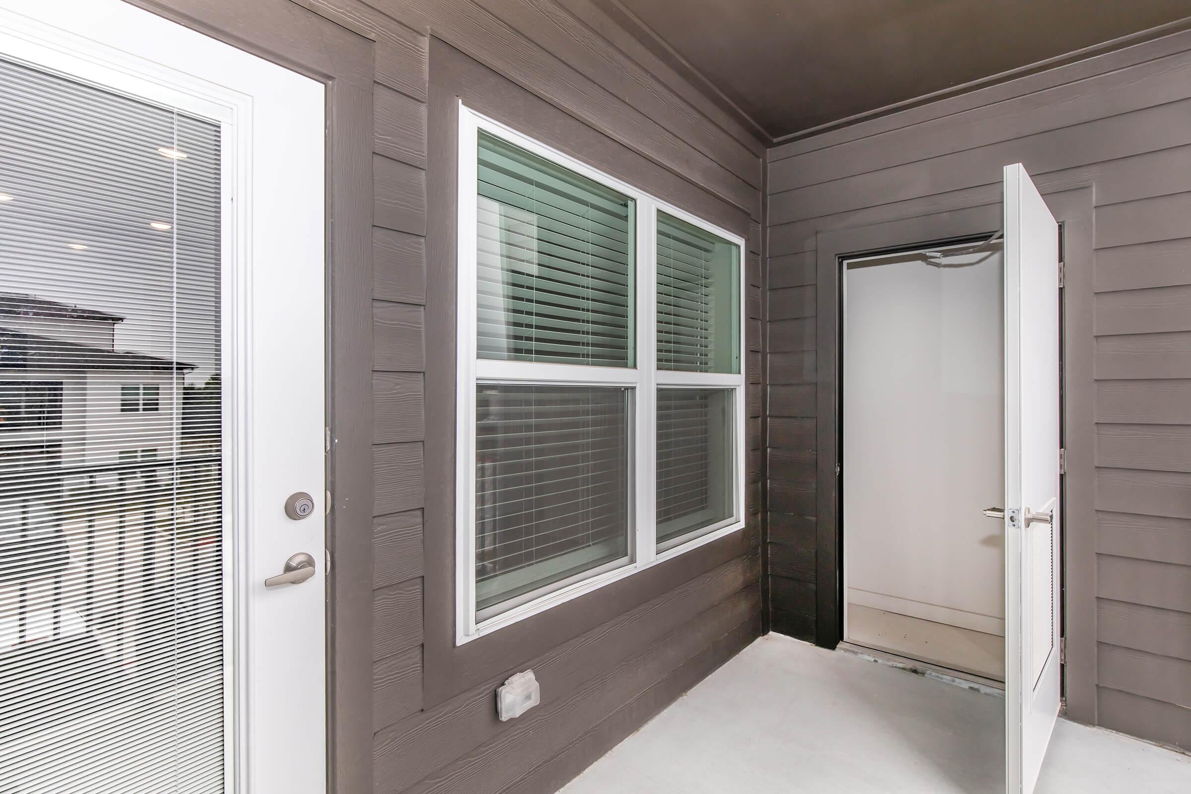 An entrance area with a modern design featuring two large windows with blinds and a partially open door leading to an interior space. The walls are painted in a dark color, and the floor is concrete. There is a separate door on the right side, suggesting access to another room or area.
