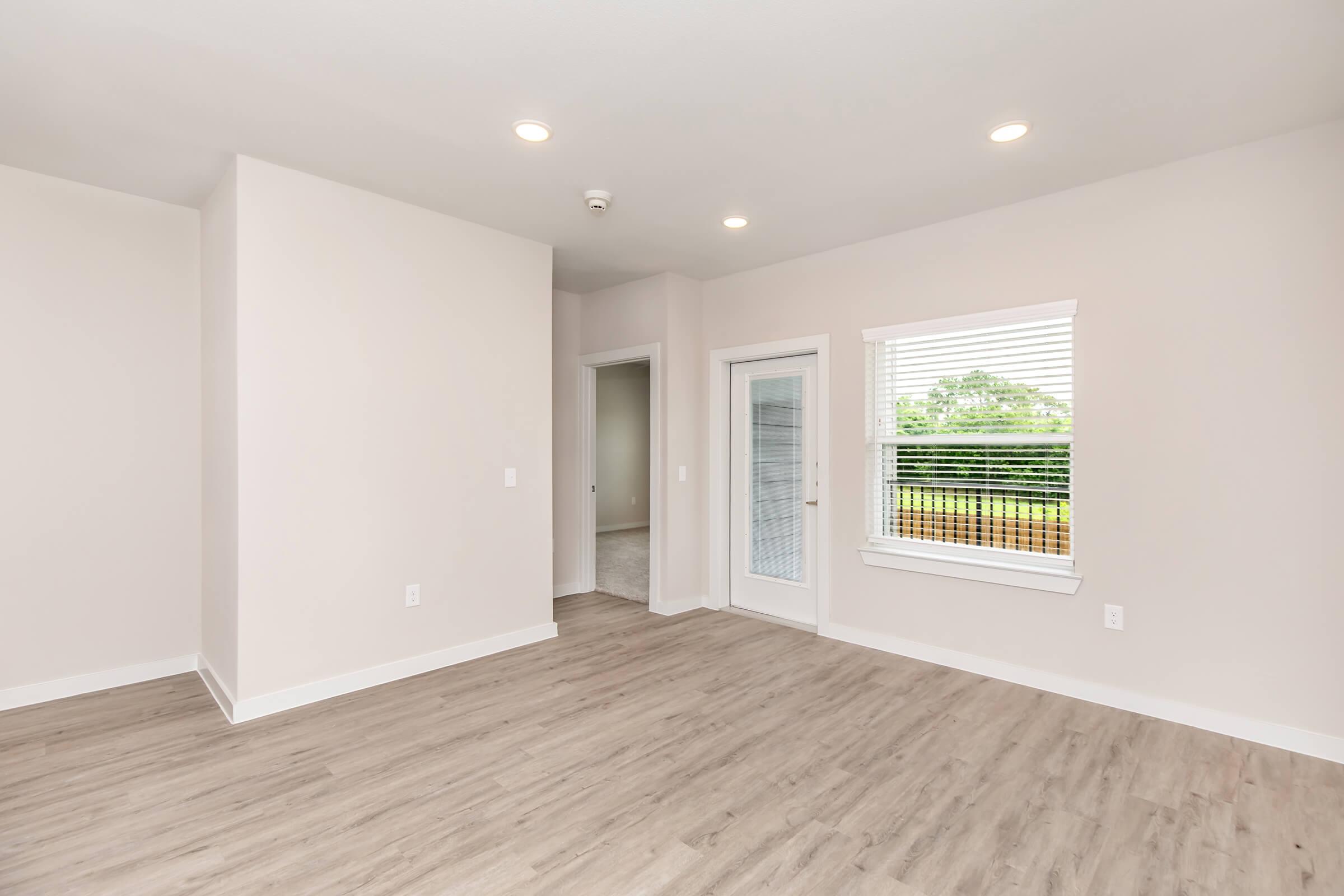 A spacious, empty room with light-colored walls and wood-like flooring. Large windows allow natural light to enter, and a door leads to an outdoor area. There is another doorway visible leading to a different room. The space appears bright and modern.