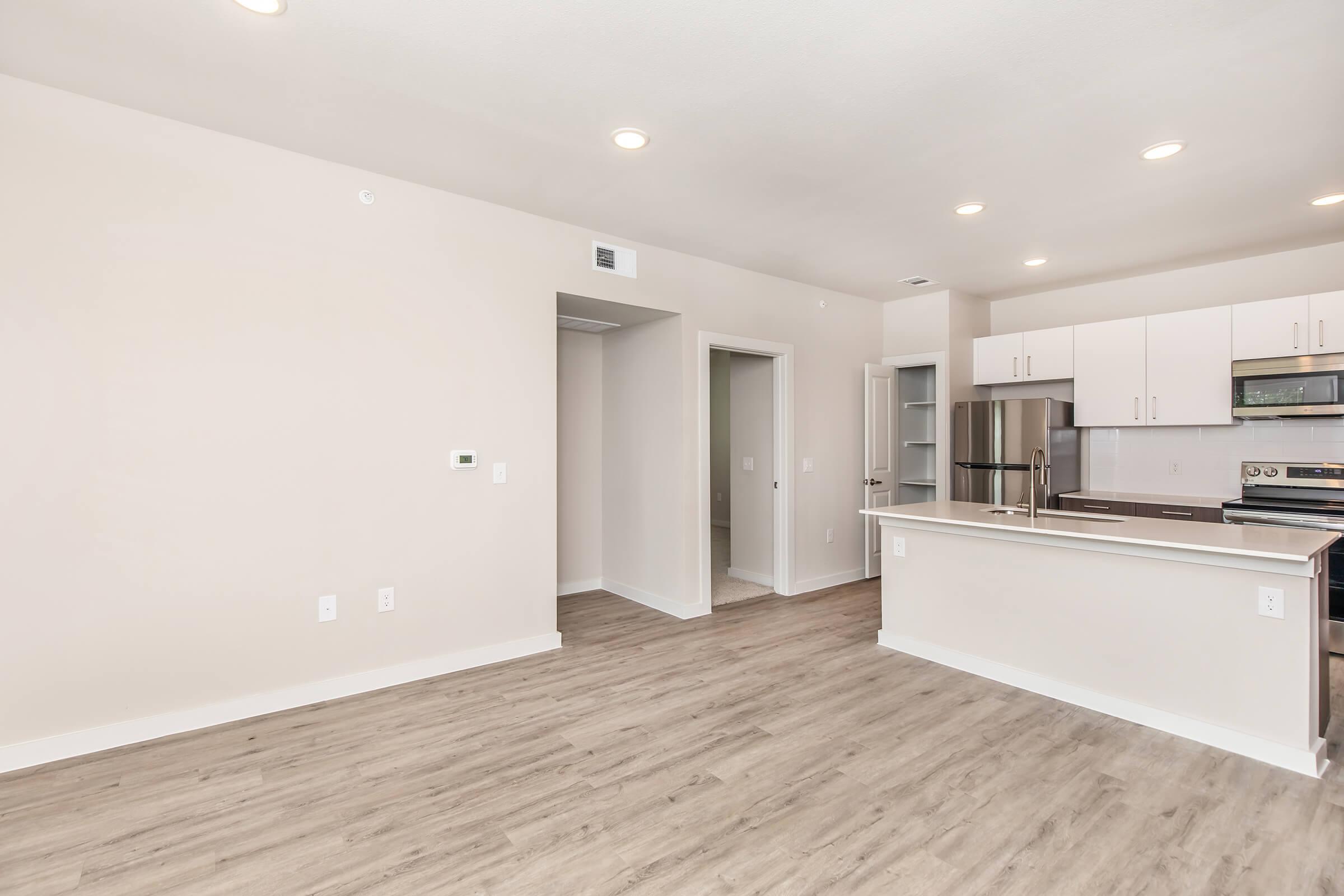 A modern, open-concept living space featuring light-colored walls and wooden floors. The kitchen is equipped with white cabinetry, stainless steel appliances, and a breakfast bar. There are doorways leading to other rooms, creating a spacious feel. Bright ceiling lights enhance the airy ambiance.