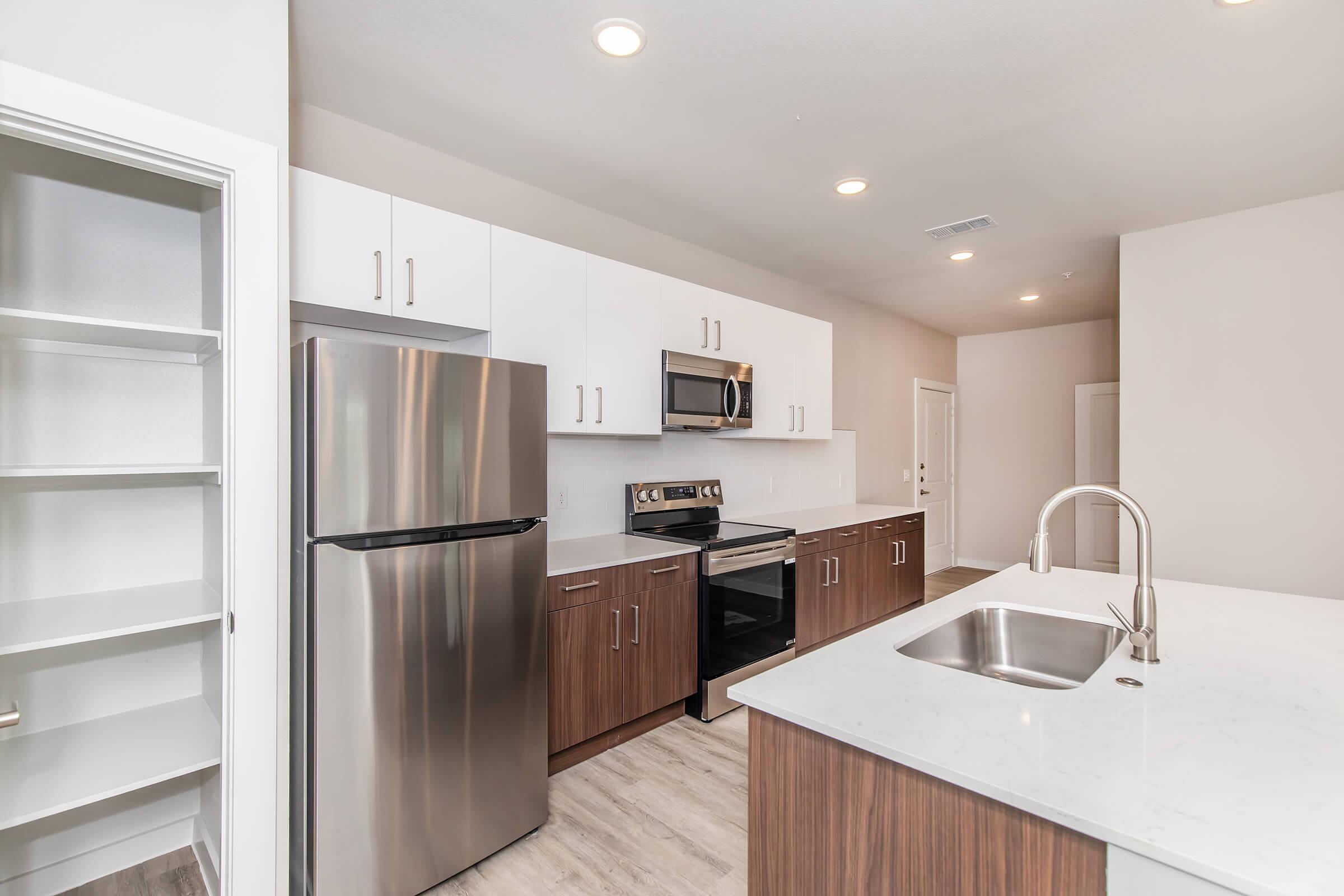Modern kitchen featuring stainless steel appliances, including a refrigerator and microwave. The cabinetry is a mix of white and dark wood, complemented by a light-colored countertop. A spacious sink is positioned on the island, with ample natural light from overhead fixtures and a bright, neutral color scheme.