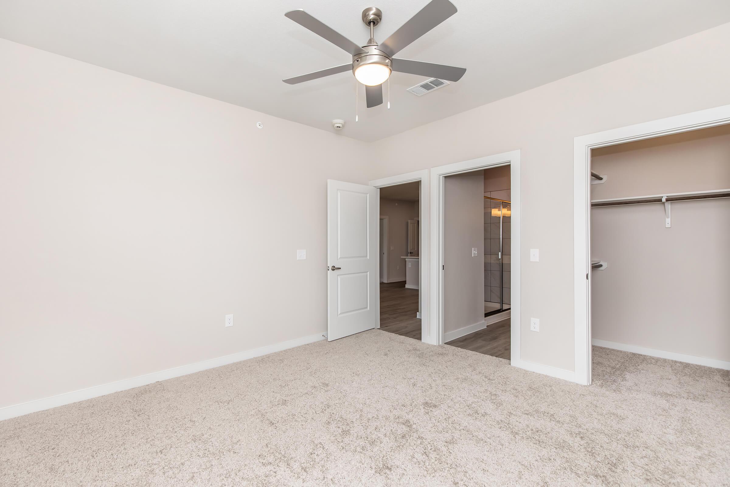 Empty room with light beige walls and carpet flooring. A ceiling fan is mounted in the center. There are two open doorways leading to adjacent rooms, and a spacious closet is visible on the right. The overall atmosphere is bright and clean, suitable for various uses.
