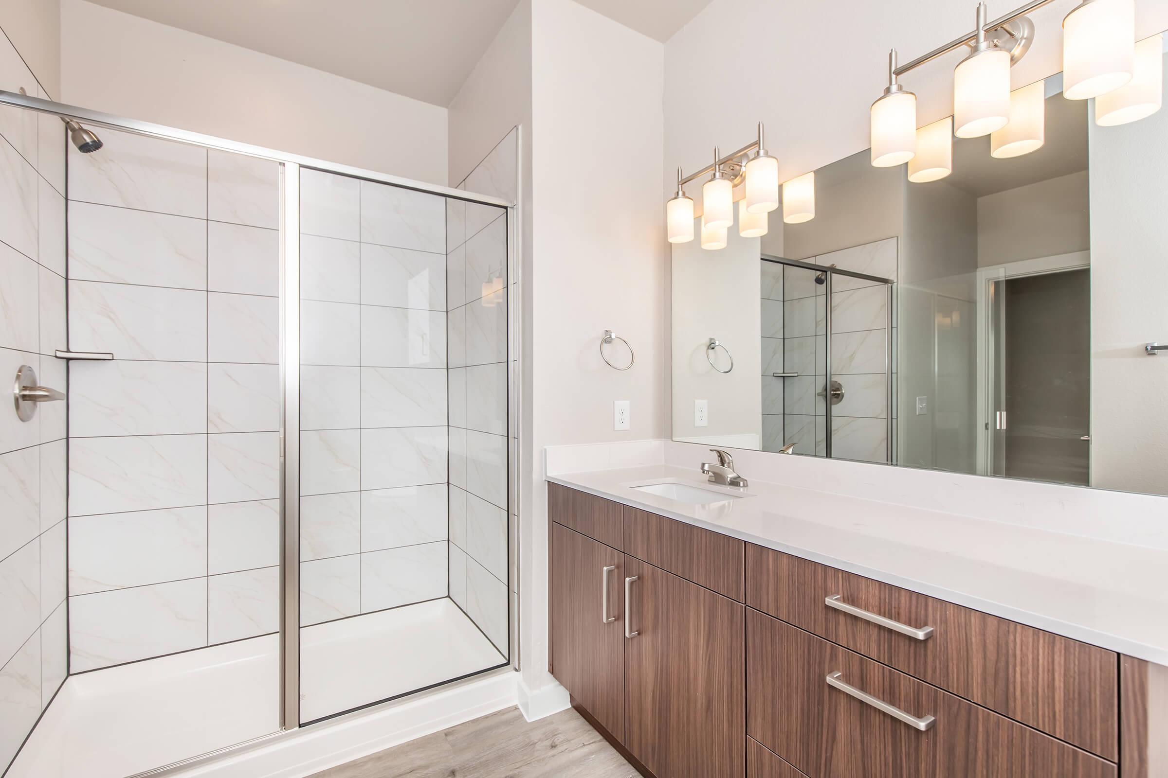 A modern bathroom featuring a glass shower enclosure, a double sink vanity with wooden cabinetry, and a large mirror with pendant lighting. The walls are painted in a light color, and the flooring appears to be a light wood finish. The overall design is clean and contemporary.