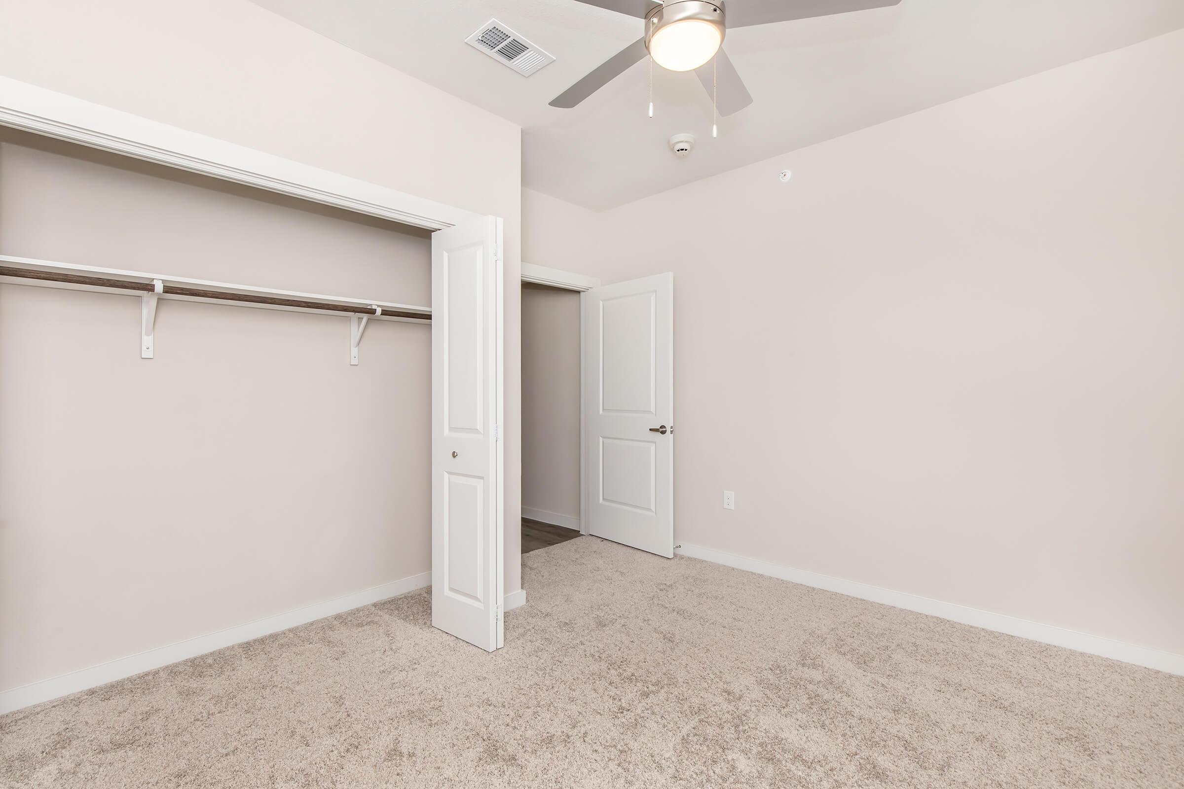 A cozy, empty bedroom featuring light beige walls and plush carpeting. The room includes a ceiling fan for ventilation, a closet with a hanging rod, and a doorway leading to another space. Natural light may be present, enhancing the inviting atmosphere.