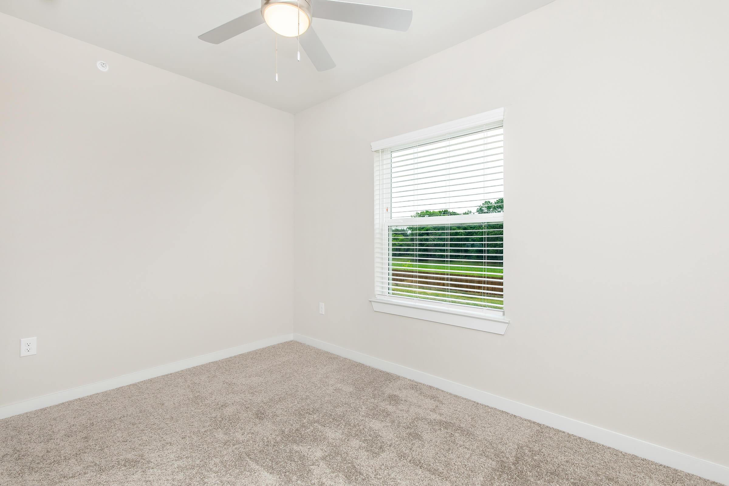 Empty room with light-colored walls and a ceiling fan. There's a window with white blinds, offering a view of greenery outside. The floor is covered with soft beige carpet, giving the space a cozy feel. The overall ambiance is bright and inviting.