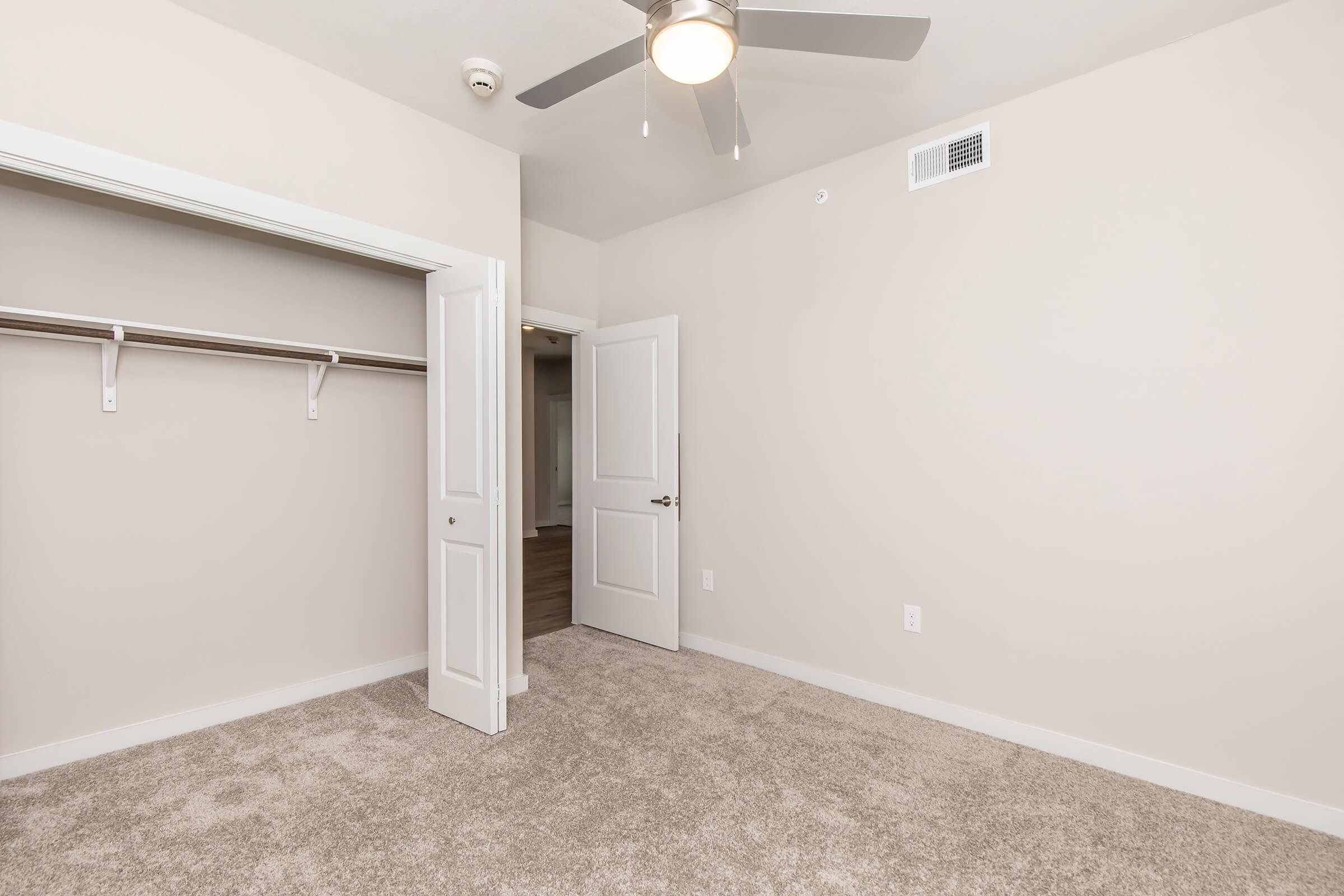 A spacious, light-colored bedroom with beige walls and carpet. It features a ceiling fan, an open closet with a single shelf, and a doorway leading to another room. Natural light illuminates the space, creating a bright and inviting atmosphere.