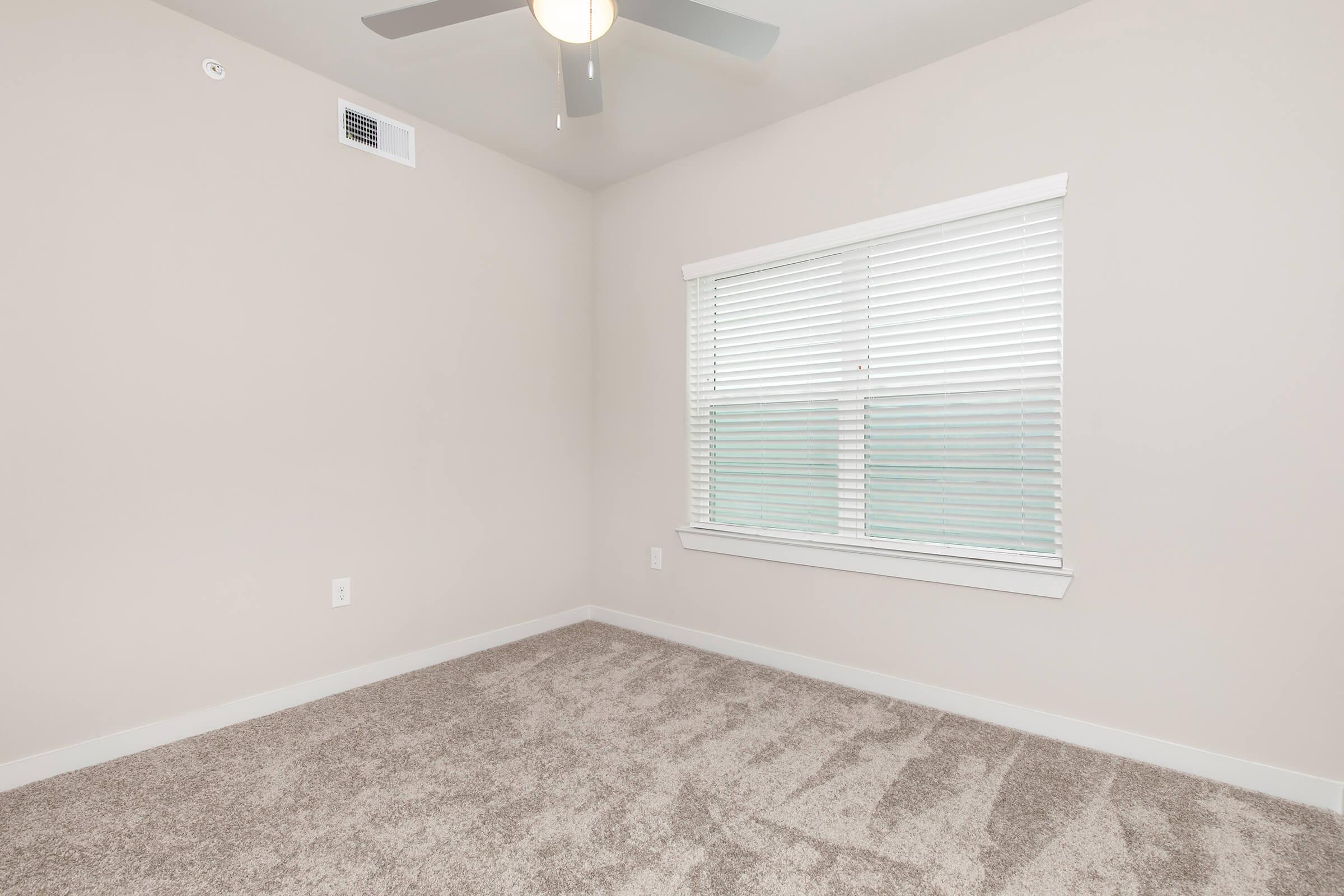 A well-lit empty bedroom with light beige walls and soft carpet. The room features a ceiling fan and a window with white blinds, allowing natural light to enter. The space is clean and uncluttered, providing a serene and inviting atmosphere.