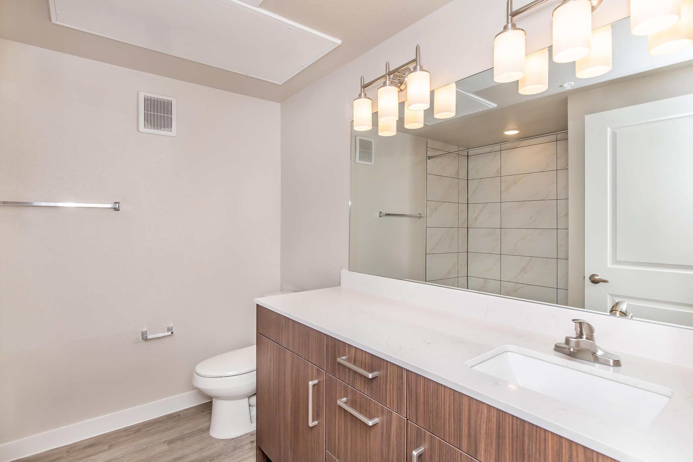 Modern bathroom featuring a white marble countertop with a sink, a wooden vanity with drawers, a contemporary mirror, and stylish overhead lighting. A toilet is located in the corner, and large tiles cover the walls. The room has a clean, bright feel with neutral colors and wood-like flooring.