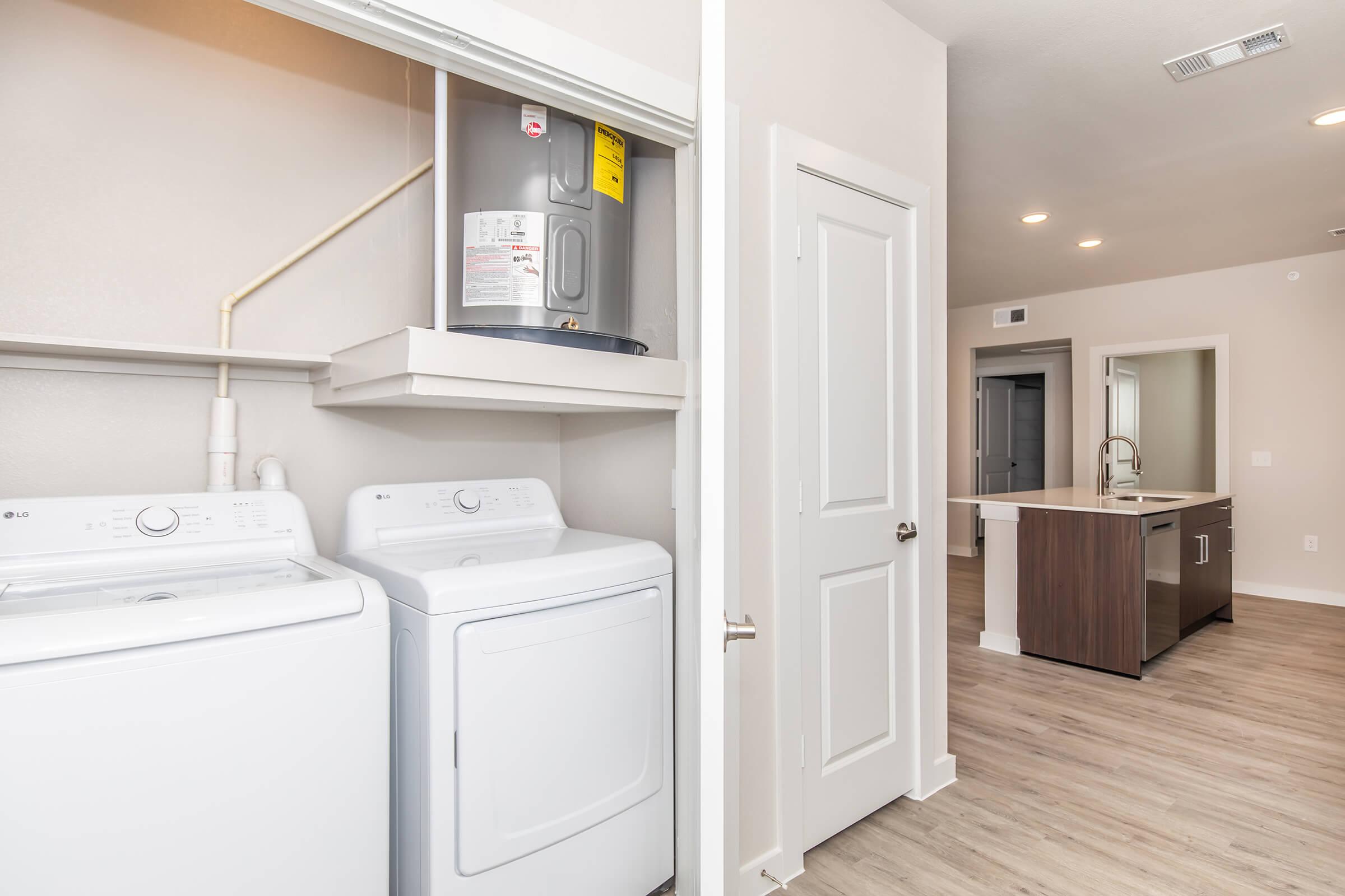 A modern laundry area featuring a stacked washer and dryer, with a hot water heater mounted on the wall. The space is bright and opens into a well-lit, contemporary kitchen area with an island and hardwood-style flooring. A door leads to another room or hallway.