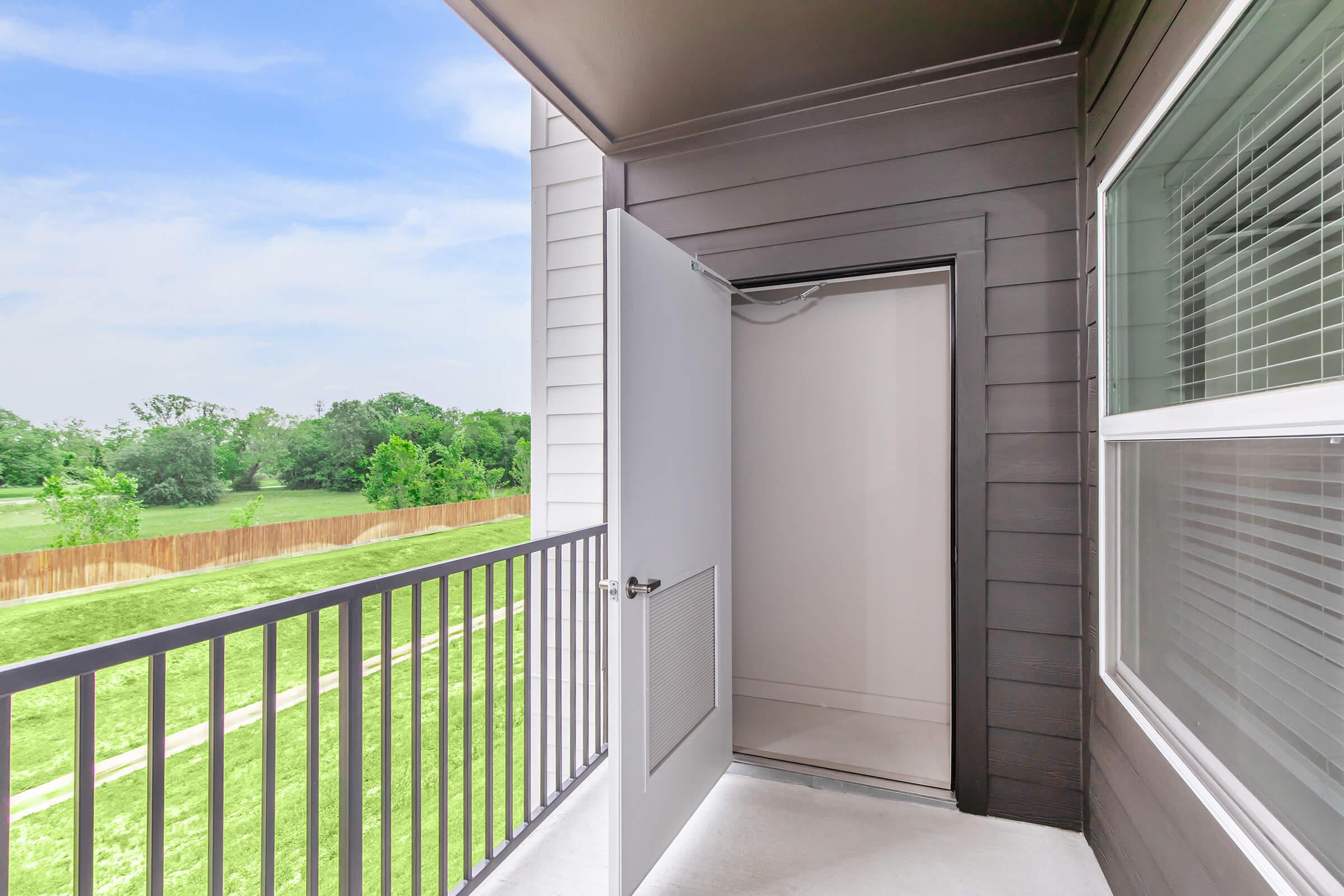 A balcony view featuring an open door leading to a small storage area. The scene includes a grassy area and trees in the background, with a clear sky above. The balcony has a light-colored railing and modern siding, creating a spacious feel.
