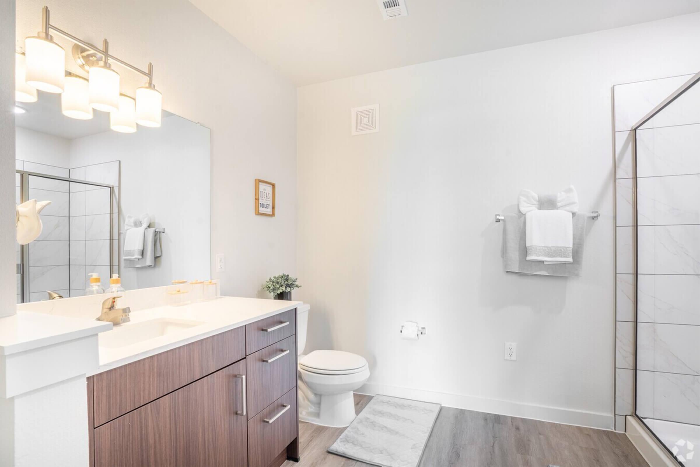 A modern bathroom featuring a large mirror above a vanity with dark wood cabinets, a white countertop, and a sink. There’s a toilet, a towel rack with white towels, and a walk-in shower with clear glass doors. The decor is minimal and bright, with neutral colors and a small plant on the counter.