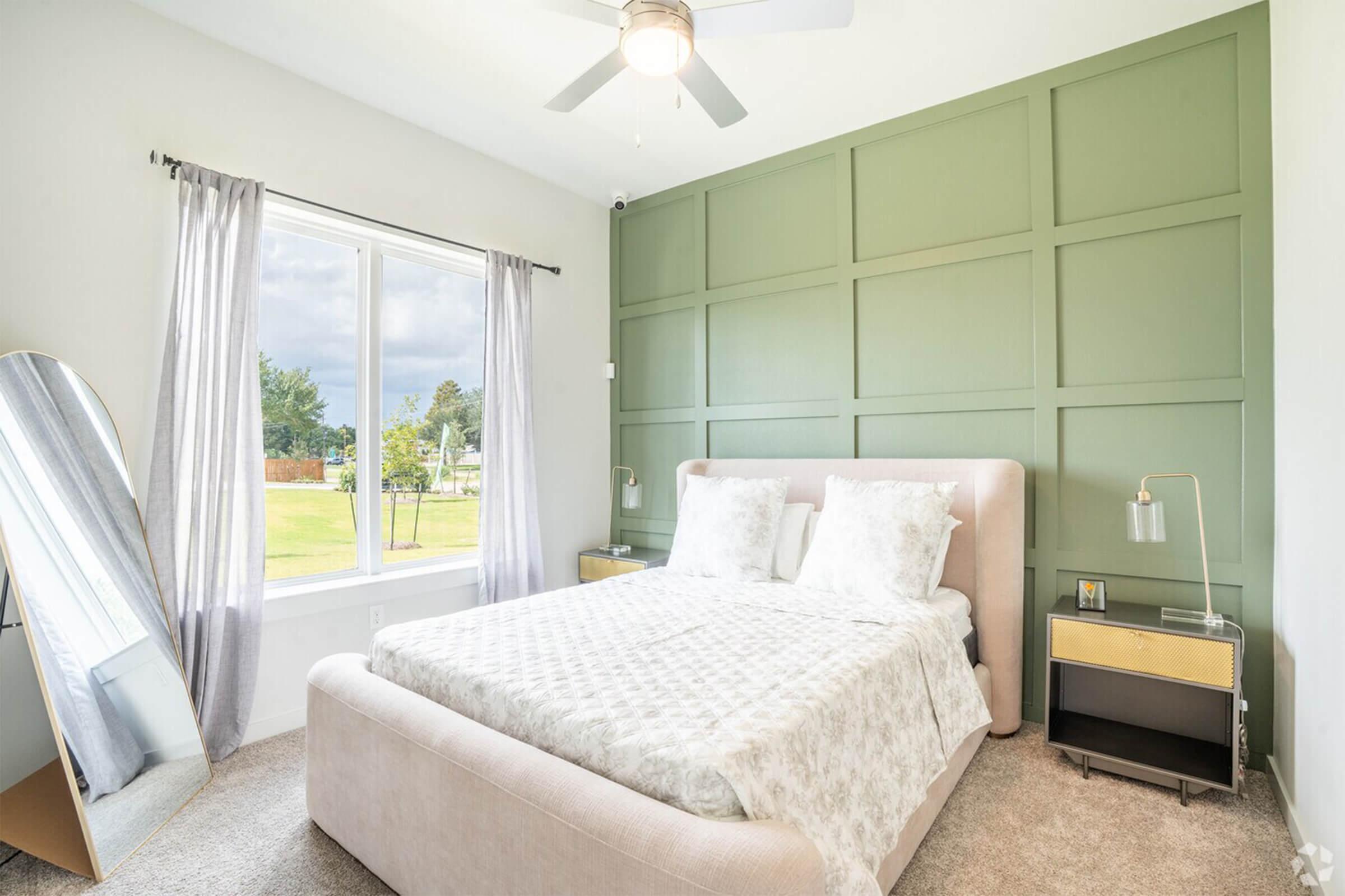 A cozy bedroom featuring a light-colored upholstered bed with decorative pillows, a stylish mirror, and two modern bedside lamps. The room has a green accent wall, large window providing natural light, and plush carpet flooring, creating a warm and inviting atmosphere.