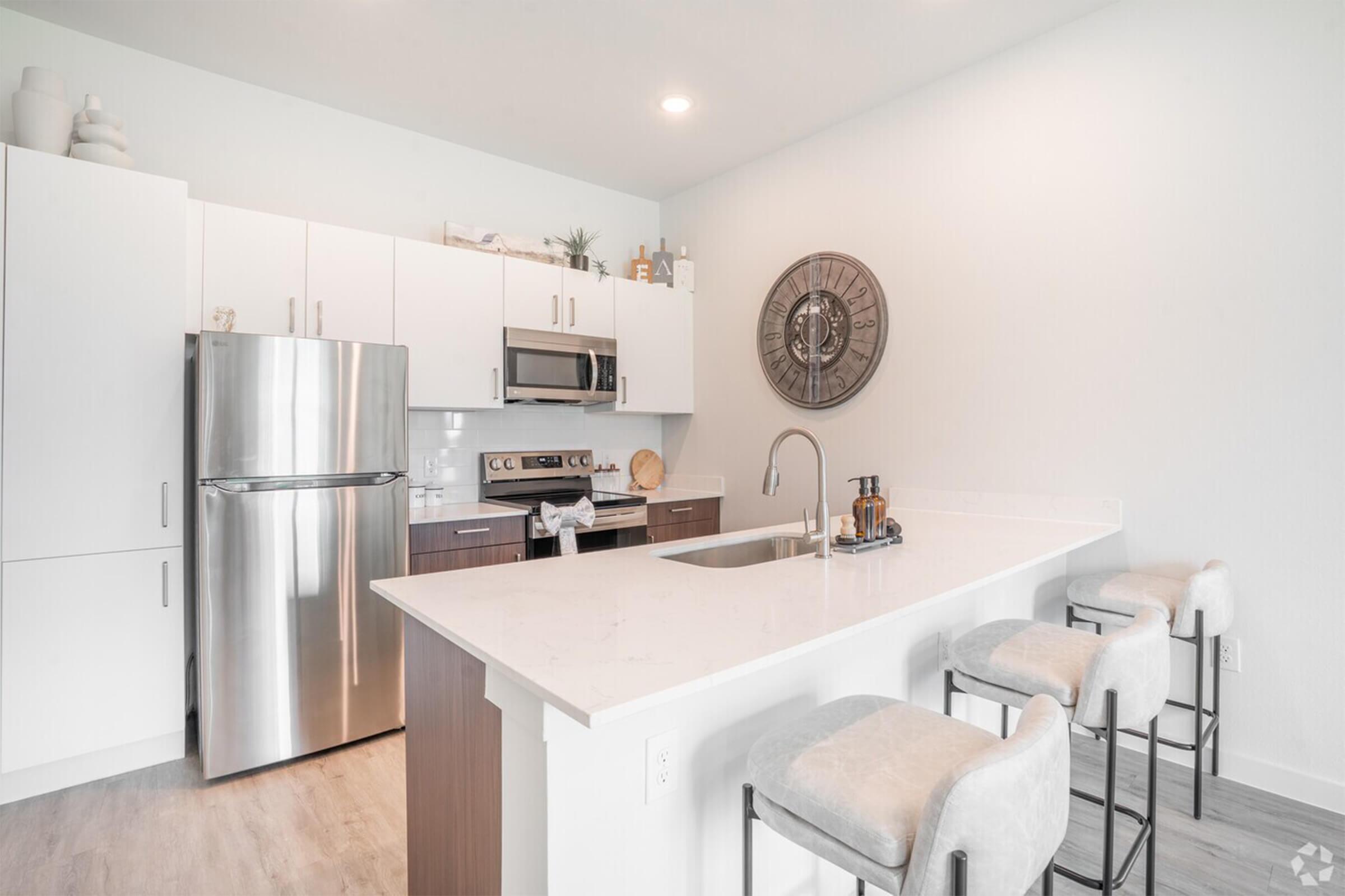 Modern kitchen featuring stainless steel appliances, including a refrigerator and microwave. A large island with bar stools offers a stylish dining area. The decor is minimalistic with light colors, wooden accents, and a circular wall clock, creating a bright and inviting atmosphere.