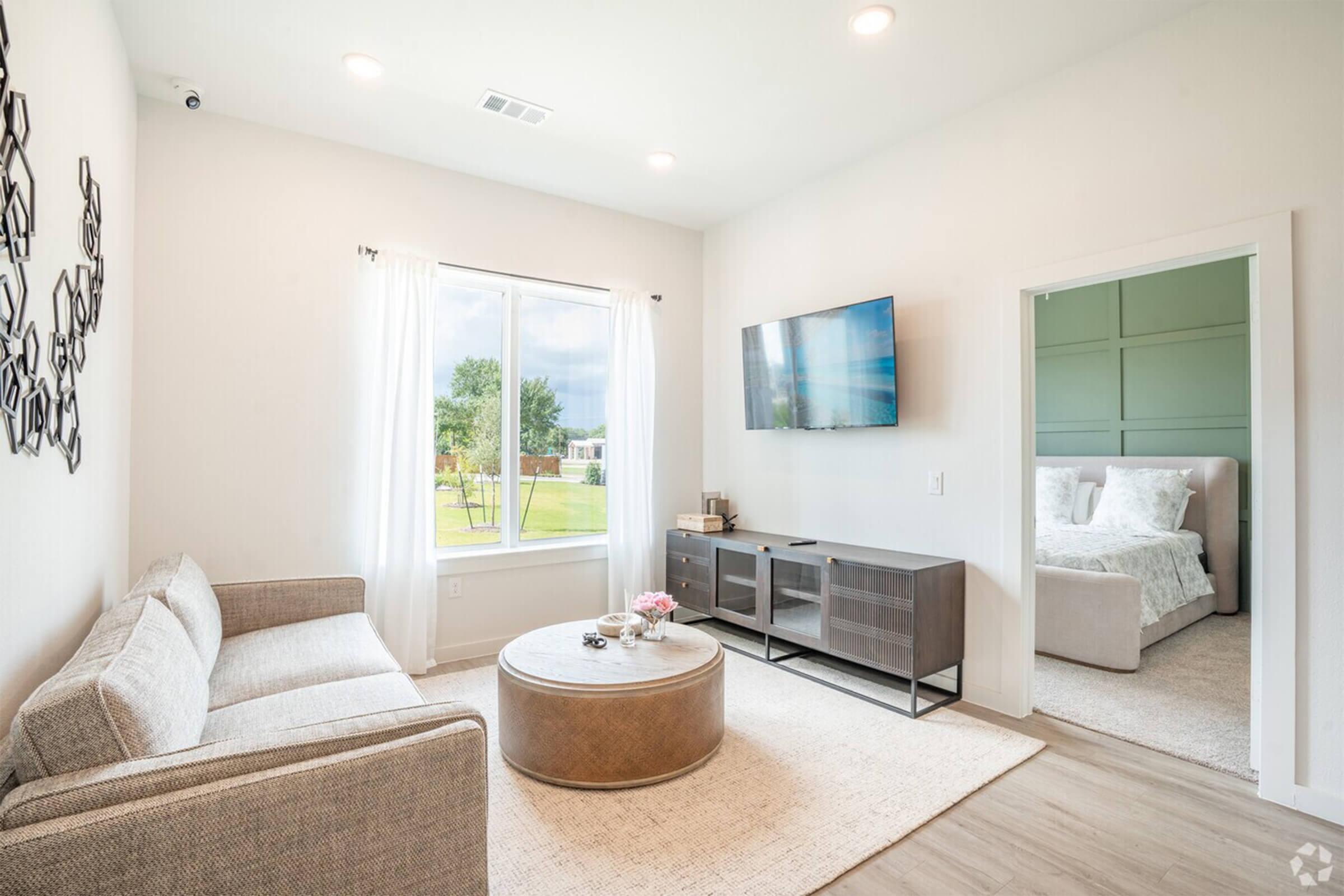 A modern living room featuring a light-colored sofa, a round coffee table, and a flat-screen TV mounted on the wall. The space has large windows allowing natural light and a view of greenery outside. A bedroom is visible through an open doorway, decorated in soft colors.