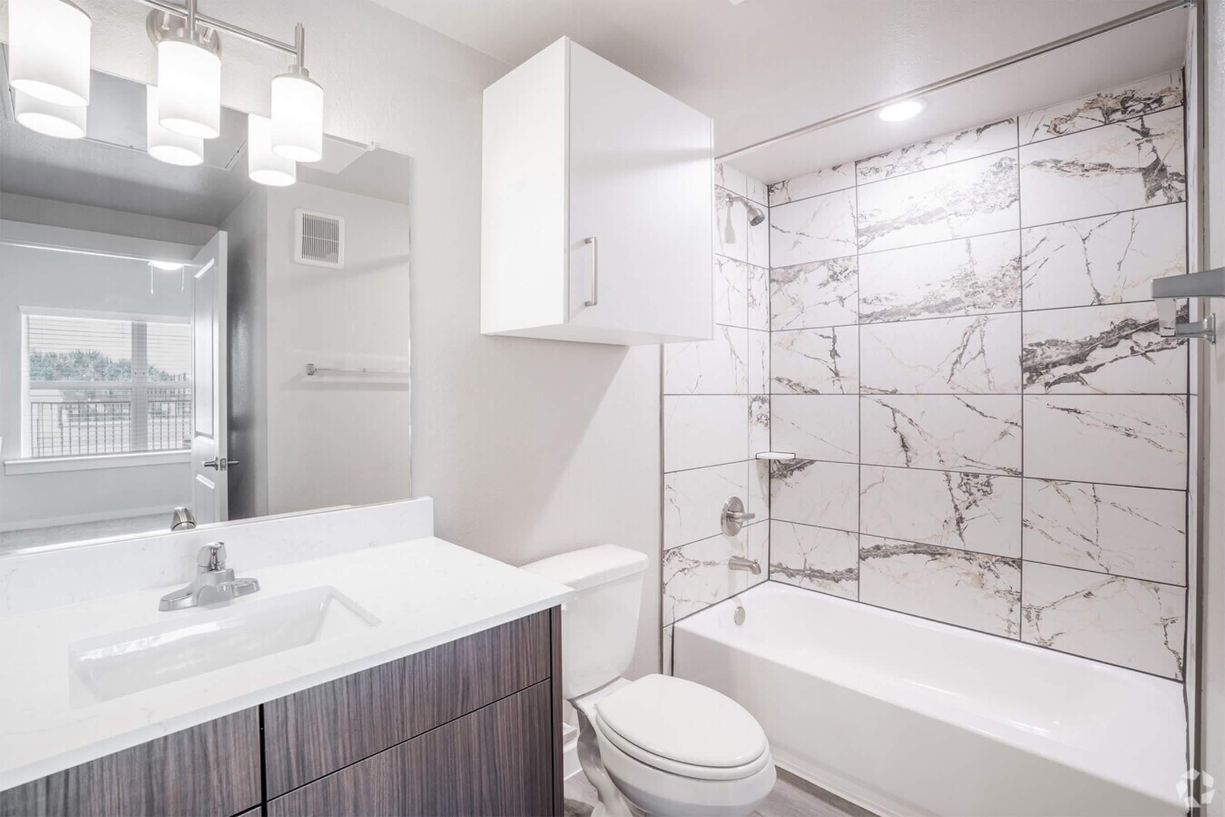 A modern bathroom featuring a bathtub with a shower, marble-patterned tiles, a floating vanity with a sink, a large mirror, and contemporary lighting fixtures. The design is sleek and minimalistic, with neutral colors and a tidy, spacious appearance. A window is visible in the background.