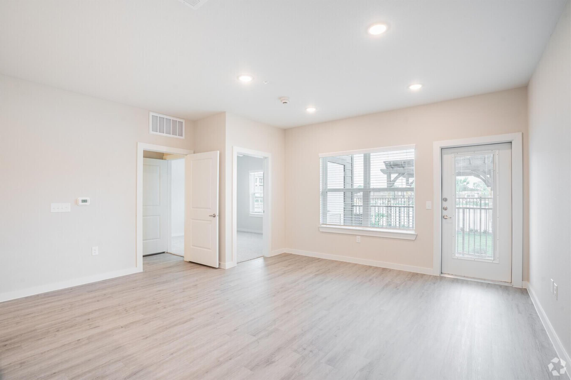 A spacious, light-filled room with light-colored walls and laminate flooring. There is a large window with a view of an outdoor area, a door leading outside, and an open doorway to another room. The space has a modern, minimalistic design, suitable for various uses.
