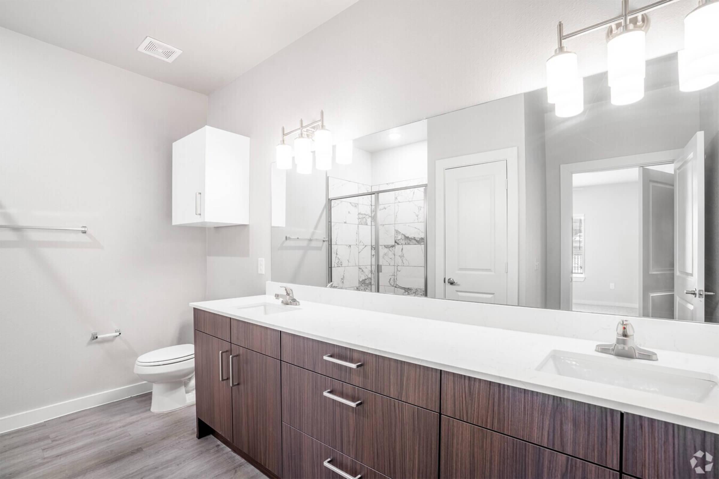 Modern bathroom featuring a double vanity with a white countertop, wooden cabinets, and two sinks. There is a spacious shower with glass doors, a white toilet, and contemporary lighting fixtures above the vanity. The walls are painted in a light tone, creating a clean and airy atmosphere.