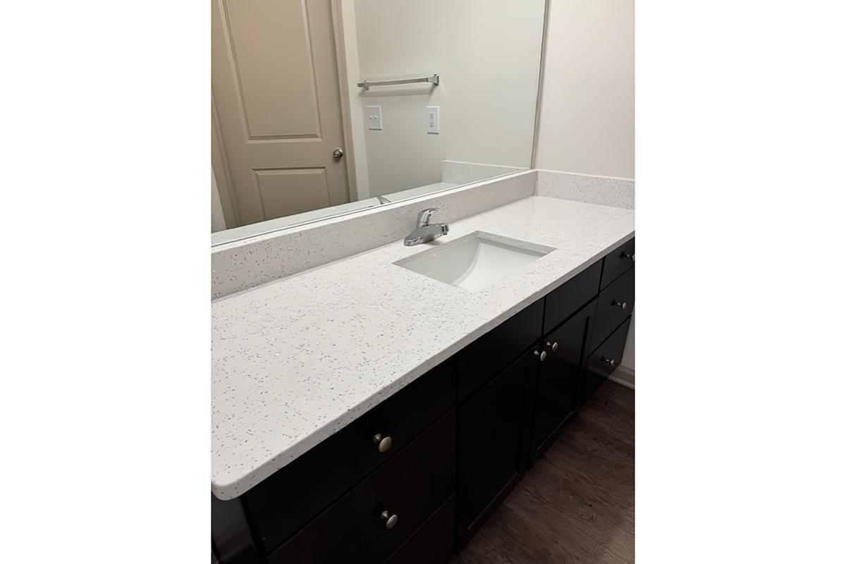 A modern bathroom countertop featuring a white speckled surface with an integrated sink. The countertop is mounted on dark wood cabinetry with several drawers and cabinets. A mirror is visible above the sink, and the bathroom has neutral-colored walls.