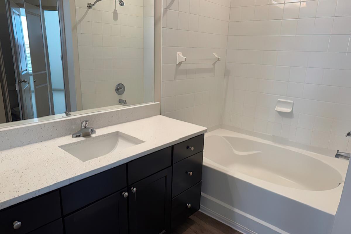 A modern bathroom featuring a white bathtub and a dark cabinetry vanity with a sink. The walls are tiled in white, and there is a large mirror above the sink. A glass shower enclosure is visible in the background. The floor appears to be made of light wood.