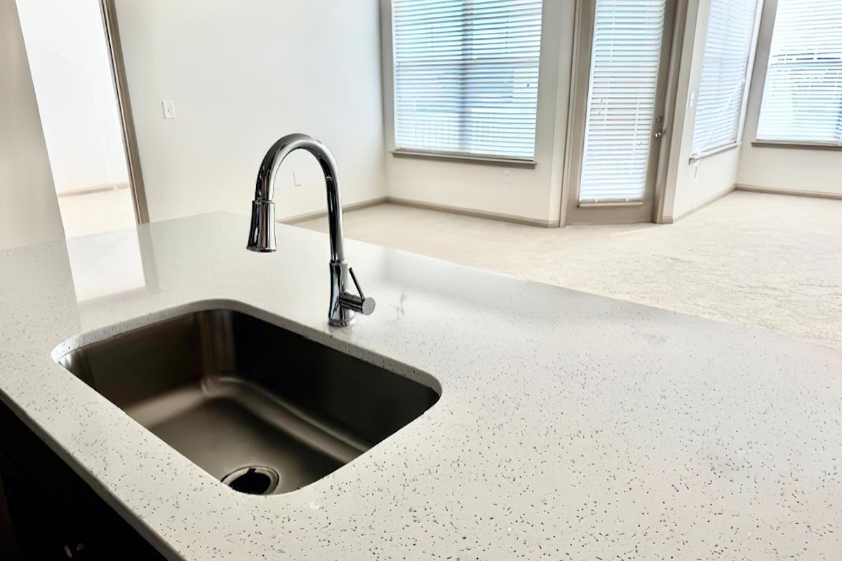 A modern kitchen countertop with a stainless steel sink and a sleek faucet. In the background, there's a spacious, well-lit living area with large windows covered by blinds, and light-colored carpeting on the floor. The overall design is contemporary and inviting.