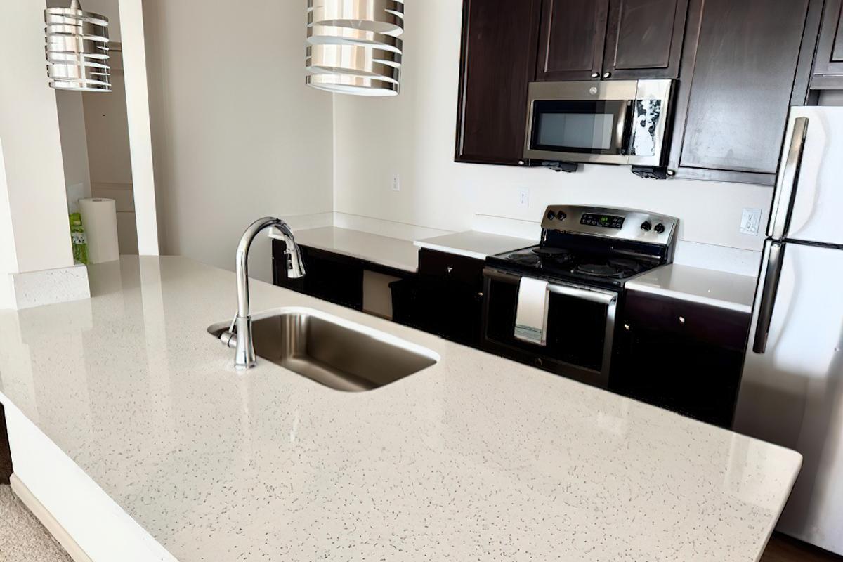 Modern kitchen with dark wooden cabinets, stainless steel appliances, and a white countertop. Features a sink with a tall faucet and pendant lighting above. The kitchen includes an oven, a microwave, and a refrigerator, creating a clean and contemporary space.