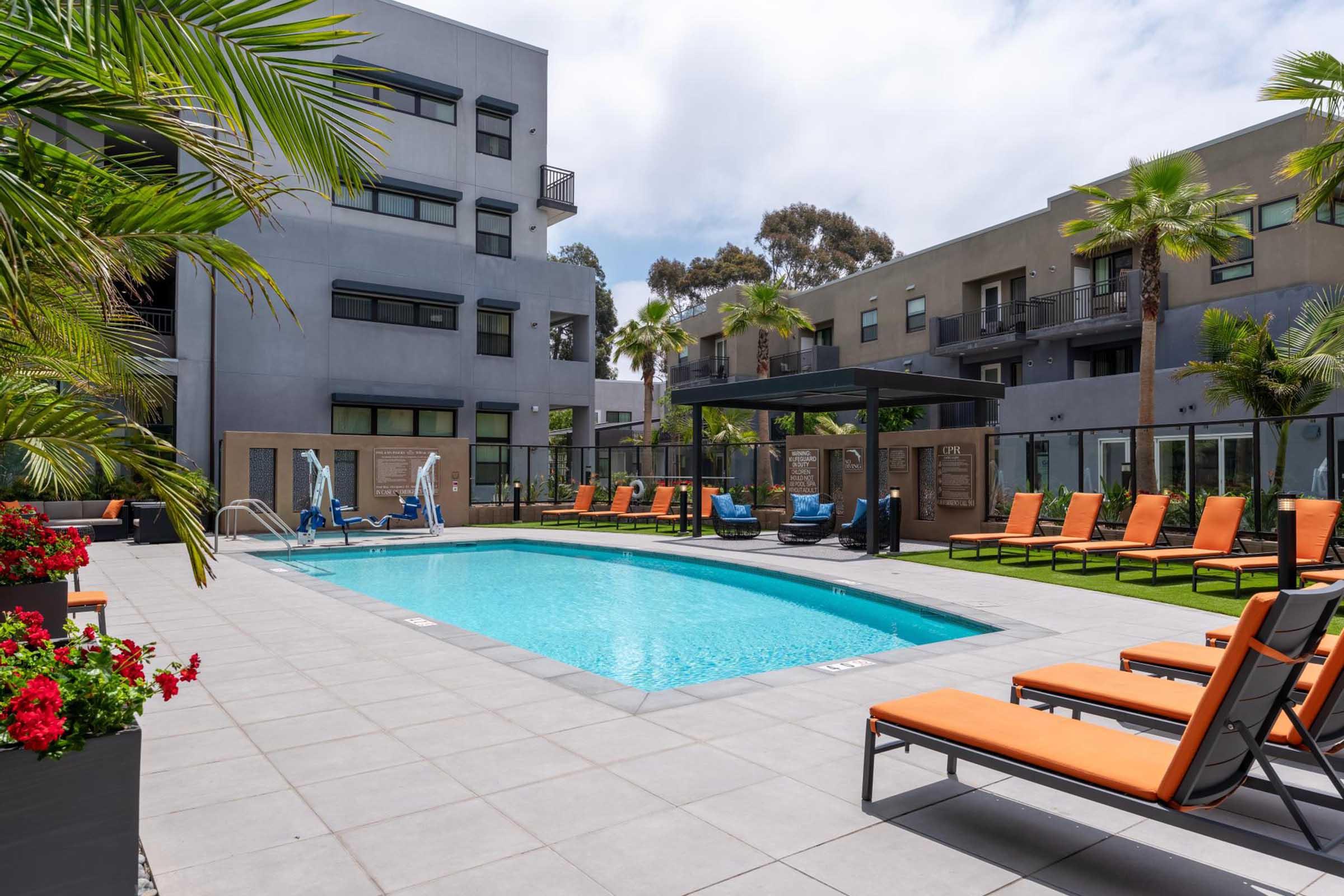 A modern outdoor swimming pool surrounded by lounge chairs with orange cushions. Palm trees and colorful flowers add greenery to the scene. Apartment buildings with large windows are visible in the background, creating a relaxed and inviting atmosphere.