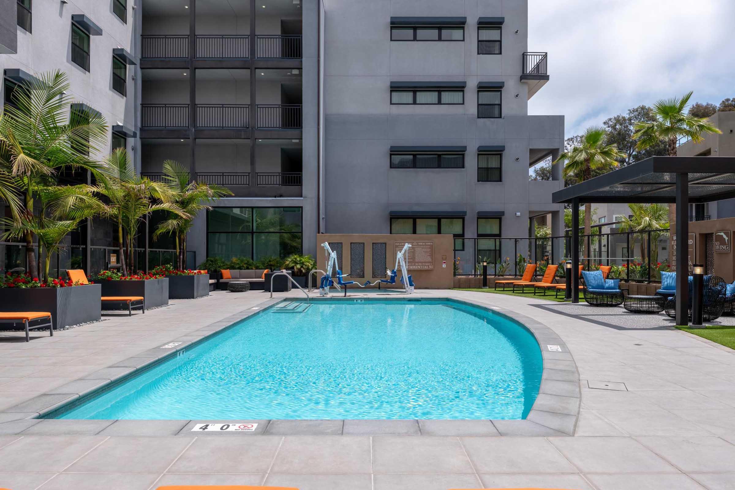 A modern swimming pool in an outdoor area surrounded by palm trees and lounge chairs. The pool is crystal clear, with nearby seating and a shaded cabana. The building in the background has multiple floors with balconies, creating a contemporary urban atmosphere.
