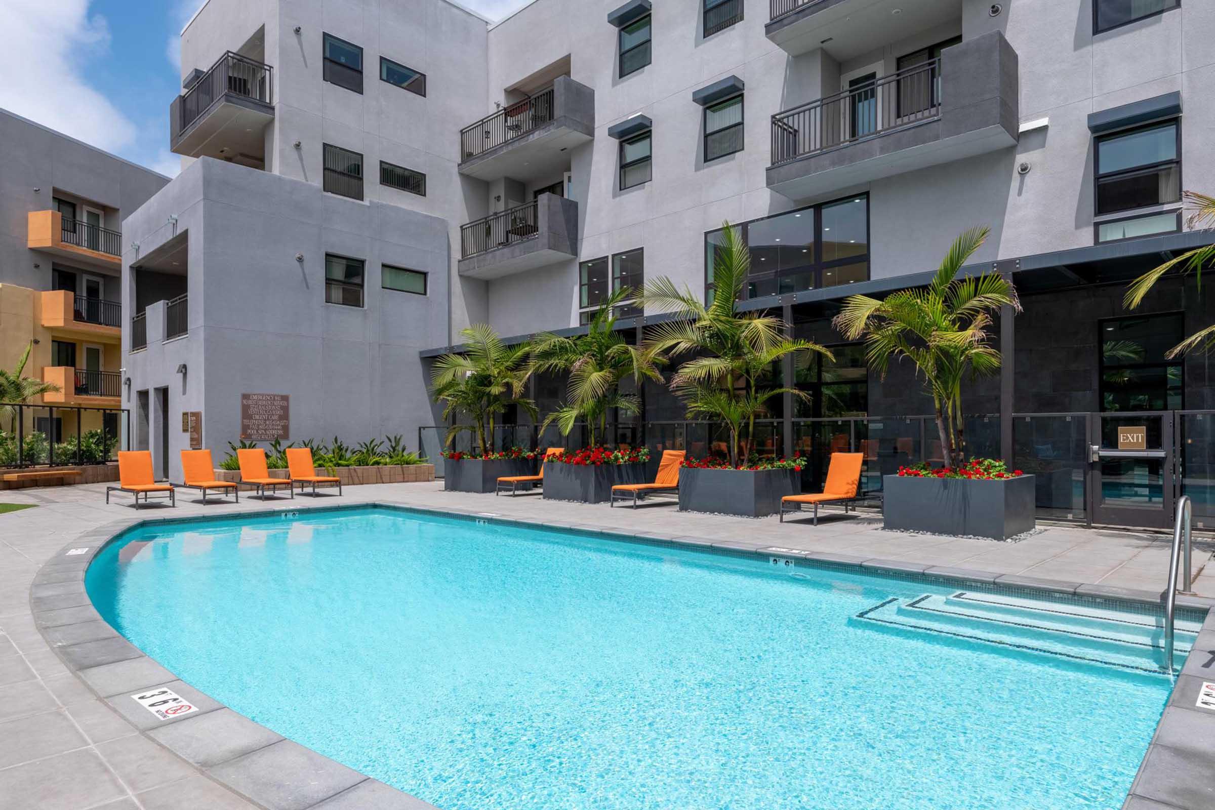 A modern apartment complex features a bright turquoise swimming pool surrounded by lounge chairs with orange cushions. Tropical plants and colorful flowers enhance the outdoor space, while the building's contemporary architecture includes large windows and balconies. Sunlight creates a vibrant atmosphere.