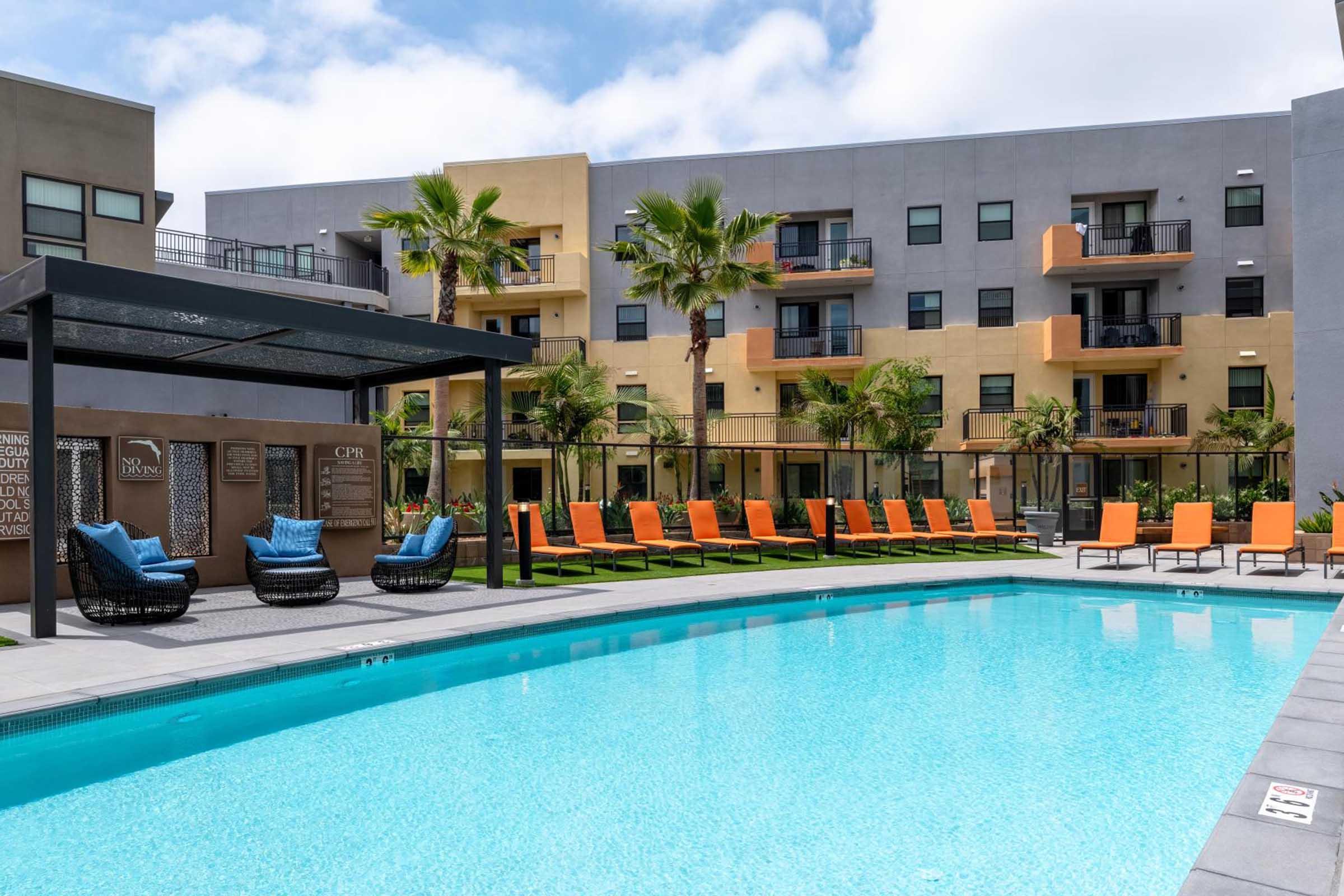 A modern apartment complex featuring a swimming pool surrounded by lounge chairs. Palm trees and green landscaping enhance the outdoor area, while buildings in the background have balconies and a contemporary design. The sky is partly cloudy, adding to the inviting atmosphere.