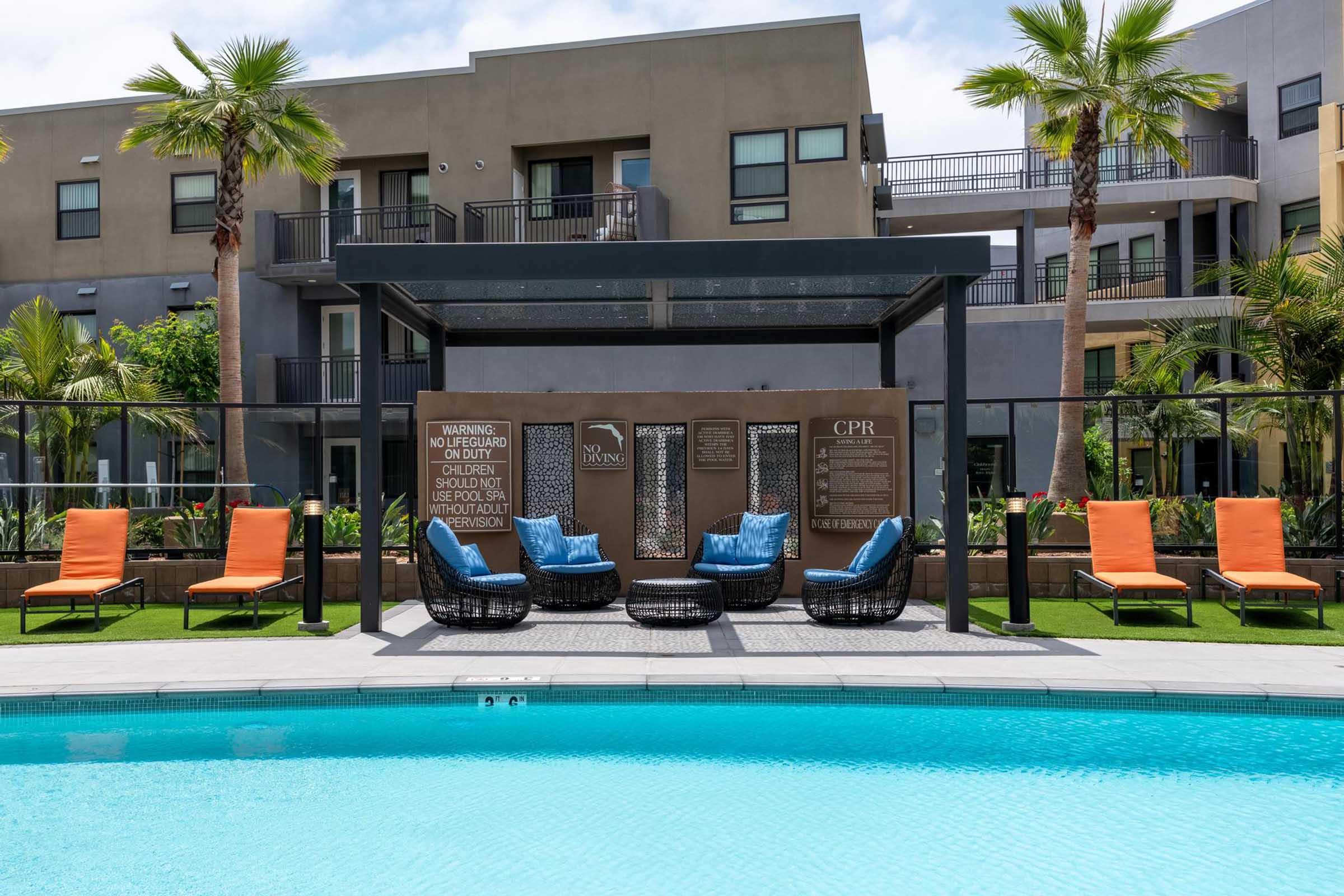 A sunny poolside area featuring blue water, lounge chairs with orange cushions, and a shaded seating area. Surrounding palm trees and modern apartment buildings create a relaxing atmosphere. Safety warning signs are displayed near the pool.