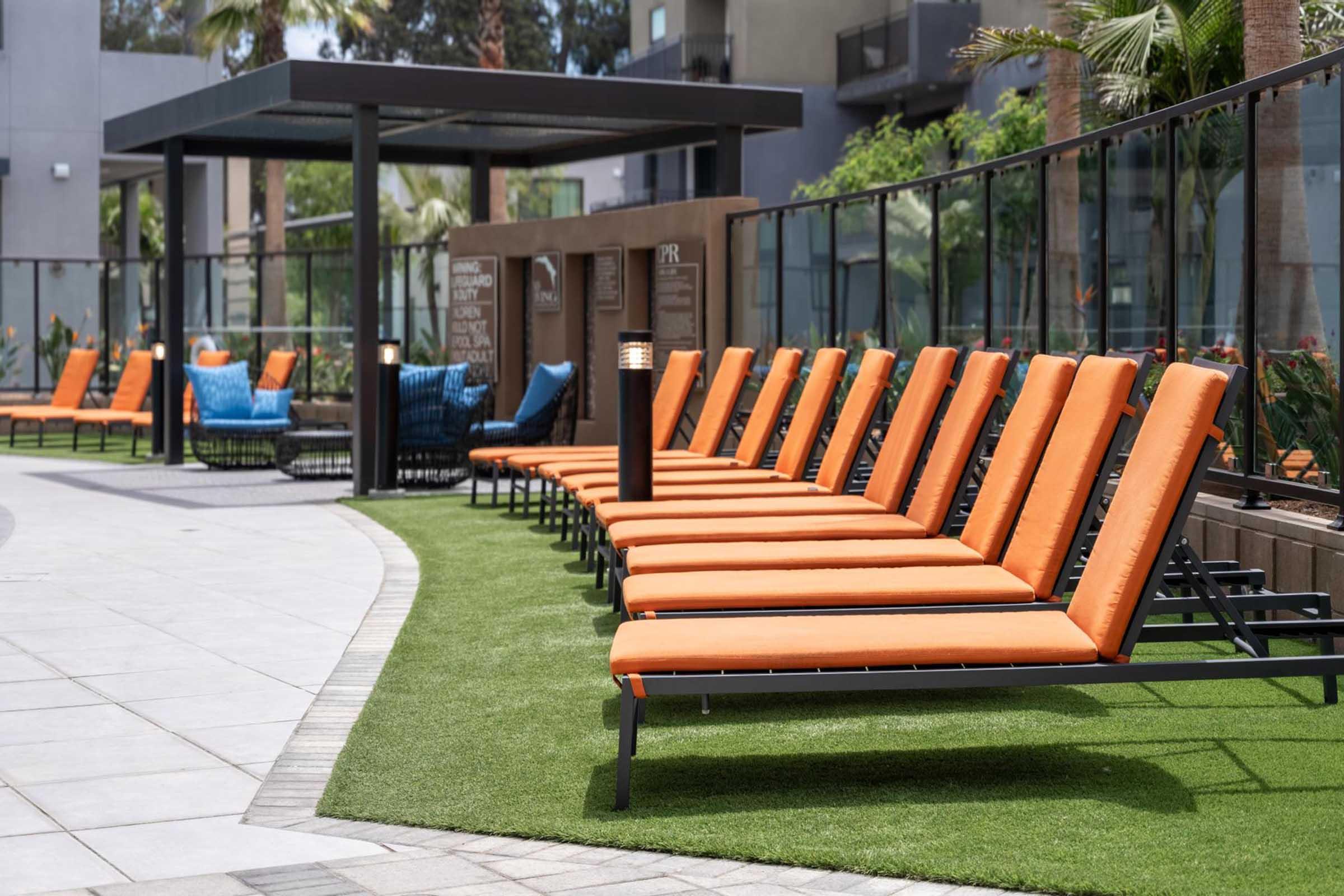Row of orange lounge chairs lined along a modern outdoor pool area, featuring artificial grass and palm trees in the background. A shaded structure is visible in the distance, creating a relaxing ambiance in a stylish setting.
