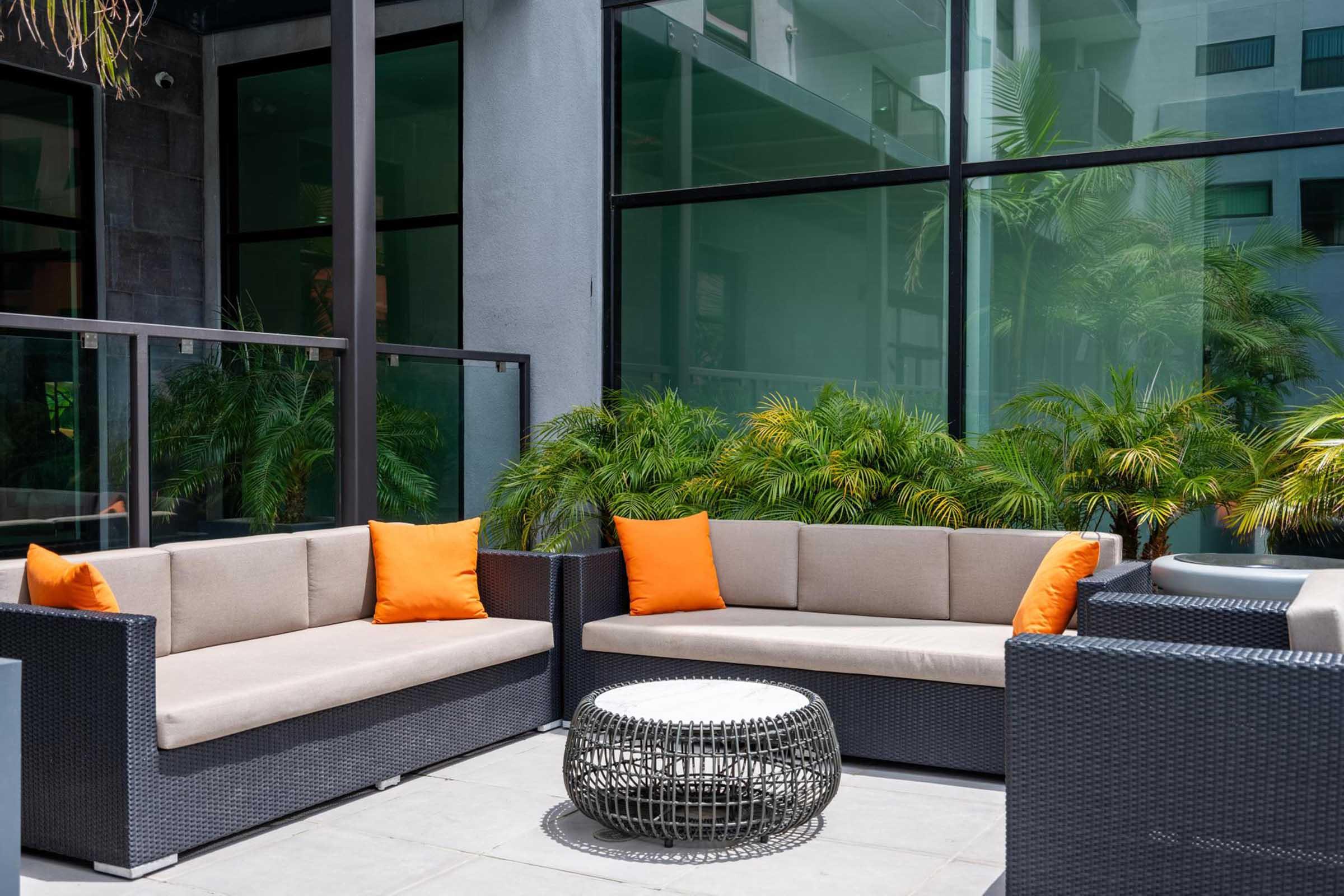 A modern outdoor seating area featuring two dark wicker sofas with light cushions and orange throw pillows, accompanied by a round woven coffee table. The backdrop consists of large glass windows and lush green plants, creating a bright and inviting atmosphere.