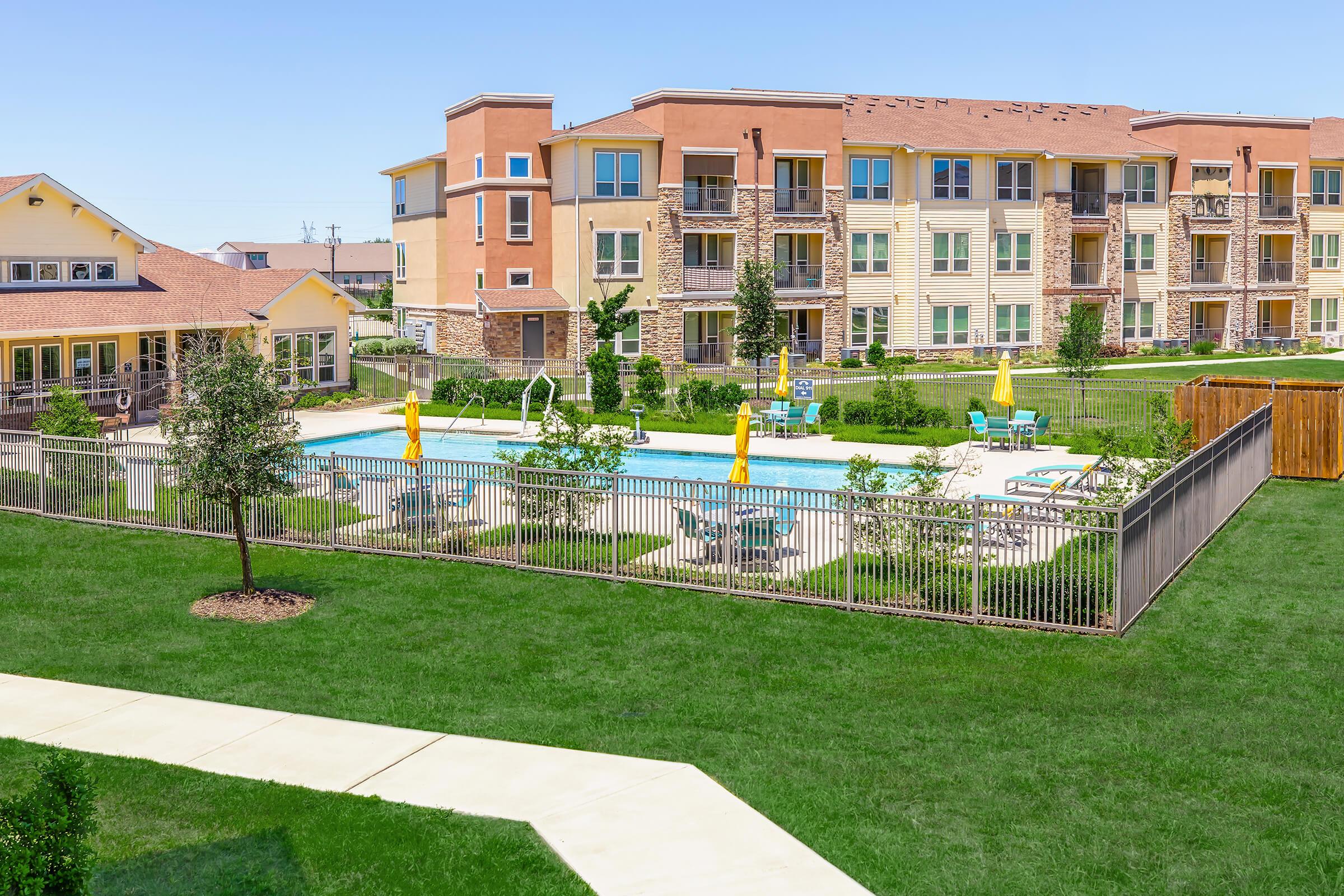 A well-maintained outdoor pool area surrounded by landscaped lawns and lounge chairs. The pool is enclosed by a fence, with several yellow umbrellas providing shade. In the background, modern multi-story apartment buildings are visible under a clear blue sky.