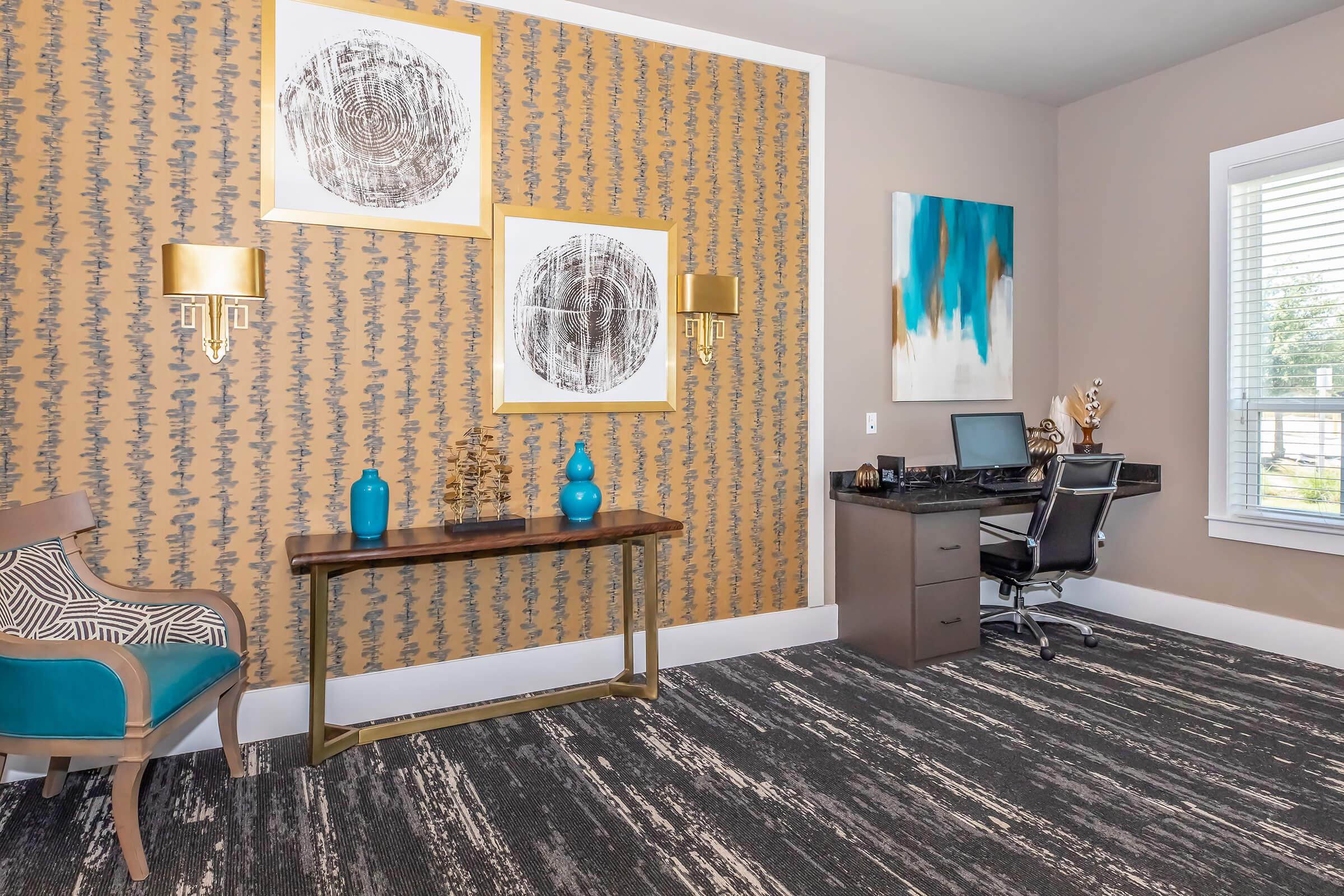 A stylish office space featuring a patterned gold and brown accent wall, two framed abstract artworks, decorative blue vases on a wooden console table, and a modern desk with an office chair. The room has a cozy feel with dark patterned carpet and natural light from a window.