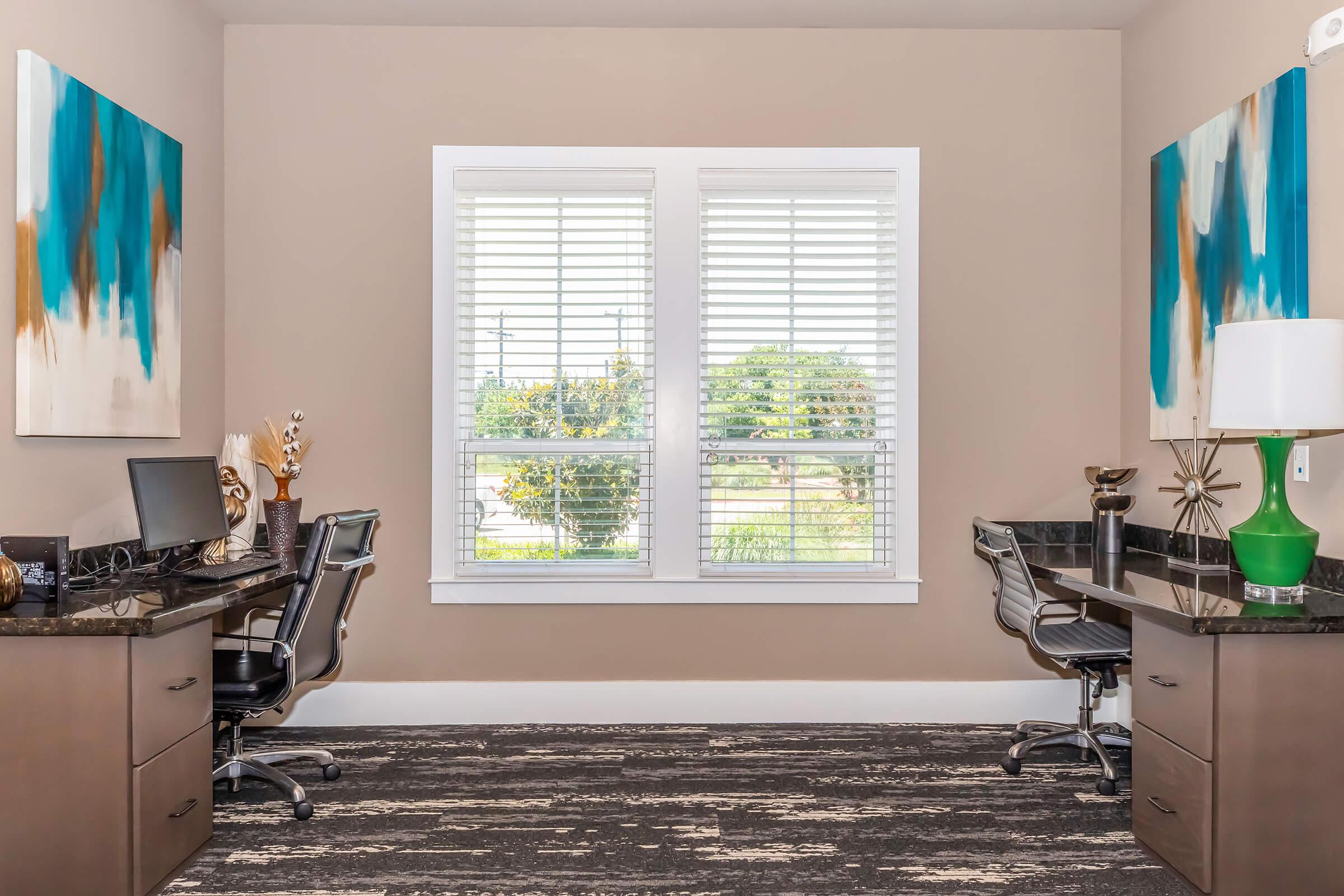 A well-lit home office featuring two workspaces with ergonomic chairs and desks. Abstract wall art in blue and gold tones adorns the walls. A large window with white blinds allows natural light to fill the space, and greenery is visible outside. The floor has a modern, patterned carpet.