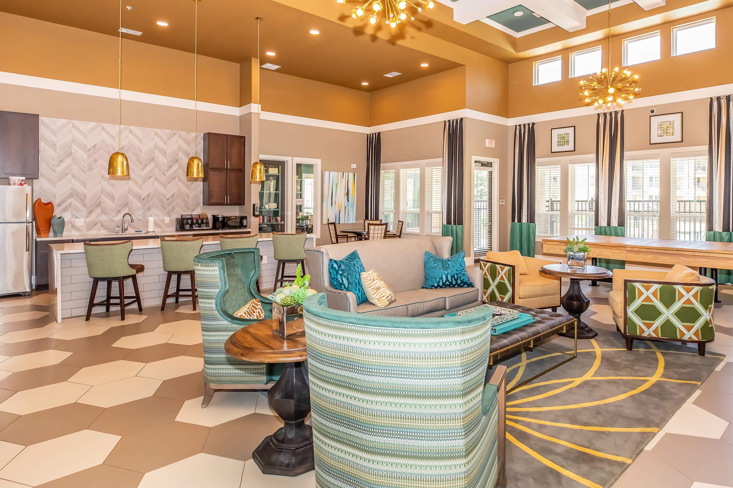 A modern communal lounge with a spacious layout featuring seating areas with stylish chairs and a large wooden table. The decor includes colorful cushions and wall art, with large windows allowing natural light. The flooring is patterned in hexagonal tiles, and there are pendant lights hanging from the ceiling.