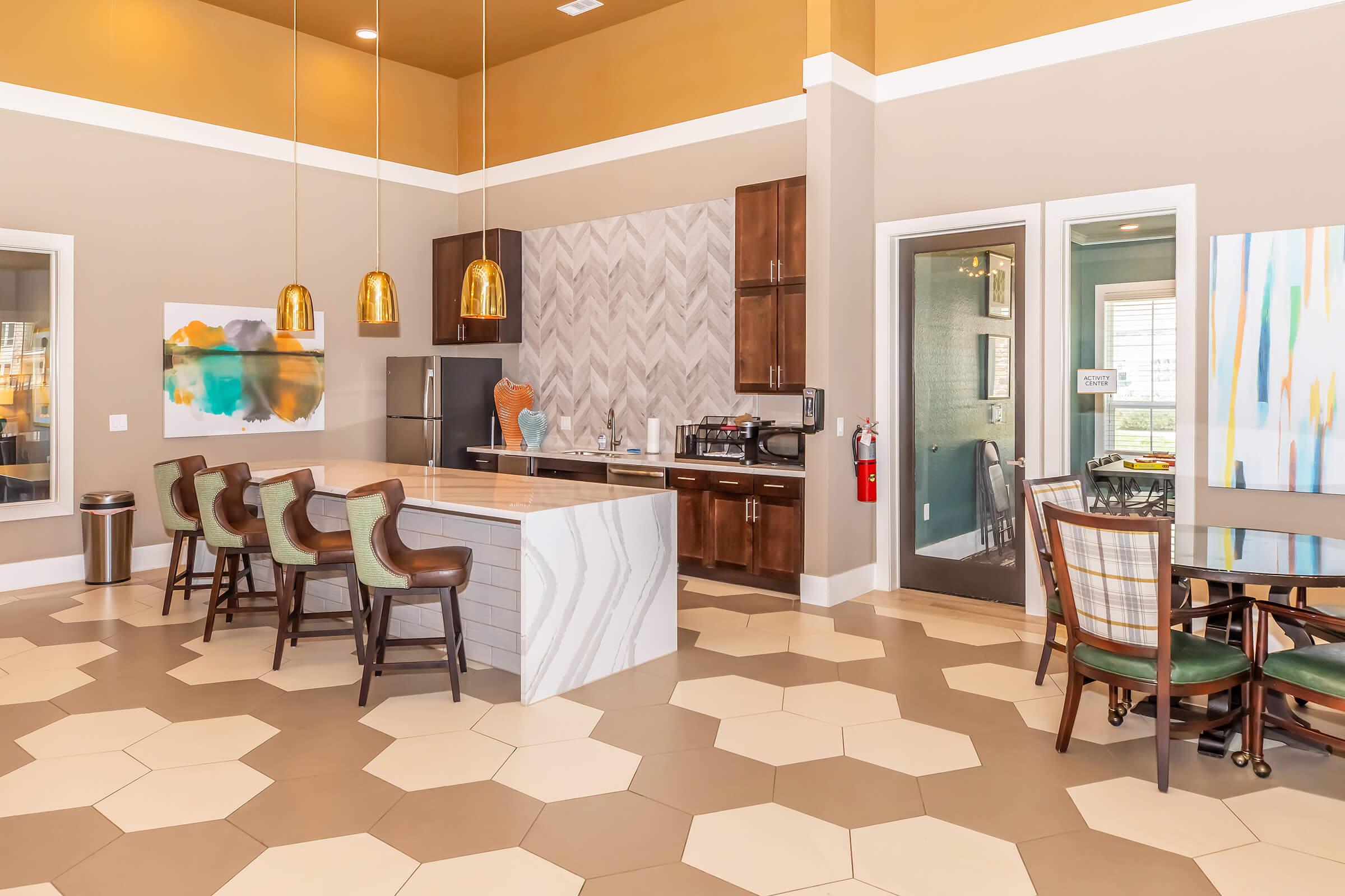 Modern kitchen and dining area featuring a large hexagonal-tiled floor, a stylish island with seating, wooden cabinetry, and contemporary light fixtures. The space includes a refrigerator, countertop appliances, and a dining table with chairs, creating a welcoming atmosphere.