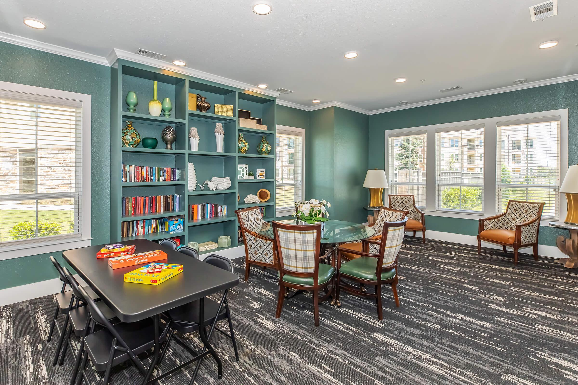 A cozy interior space featuring a reading nook with a bookshelf filled with books, a dining table and chairs, and two comfortable armchairs. The walls are painted a calming teal, and there is natural light coming through large windows. A game is set on the dining table, adding a cheerful touch.