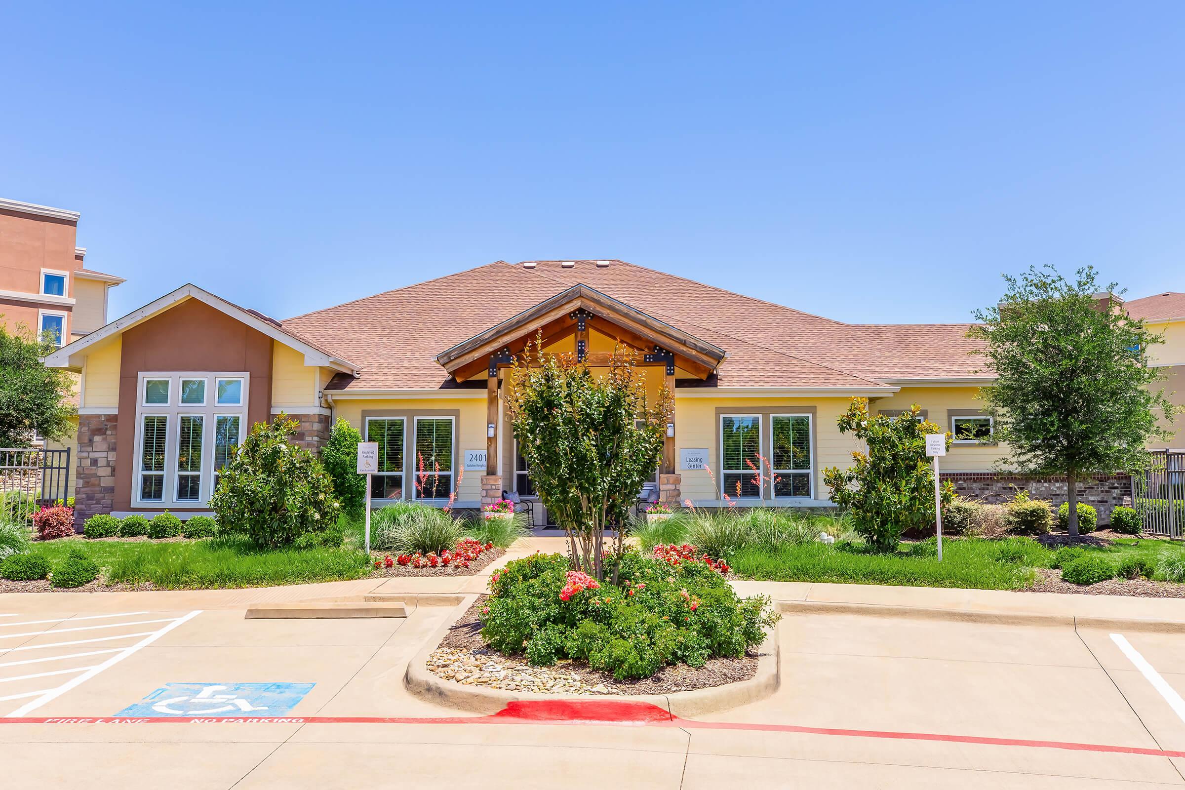A well-maintained building with a charming architectural design, featuring a sloped roof and wooden accents. The exterior includes large windows, landscaped gardens with colorful flowers, and parking spaces. A clear blue sky enhances the inviting atmosphere of the property.