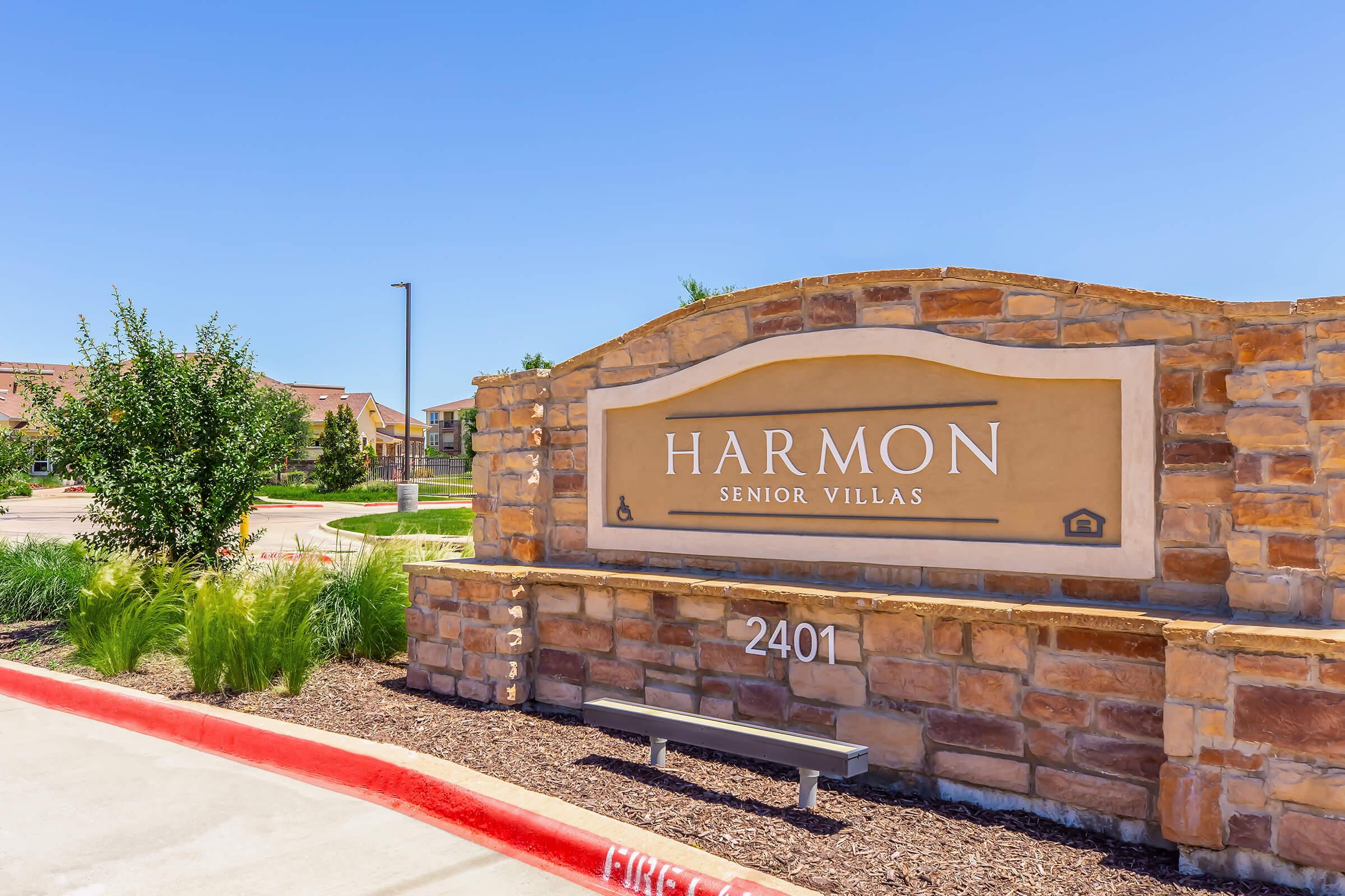 Sign for "Harmon Senior Villas," featuring a stone and concrete design with landscaping behind. The sign is prominently displayed under a clear blue sky, indicating the entrance to a senior living community. Address noted: 2401.