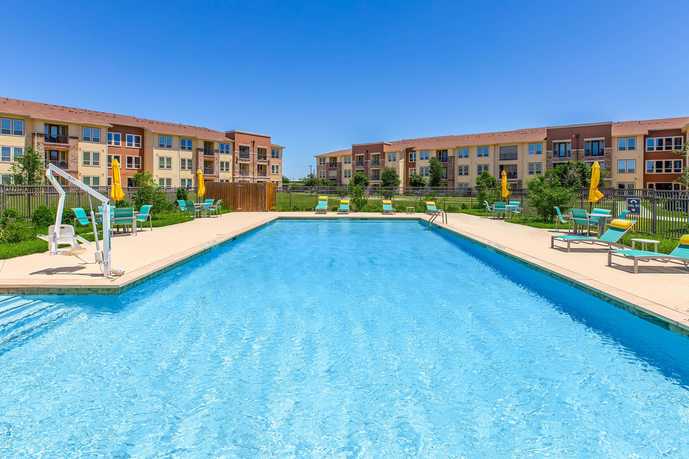 A clear blue swimming pool surrounded by lounge chairs and umbrellas, with modern apartment buildings in the background. The sky is bright blue, and the area is neatly landscaped with grass and trees, creating a welcoming outdoor space for relaxation and leisure.