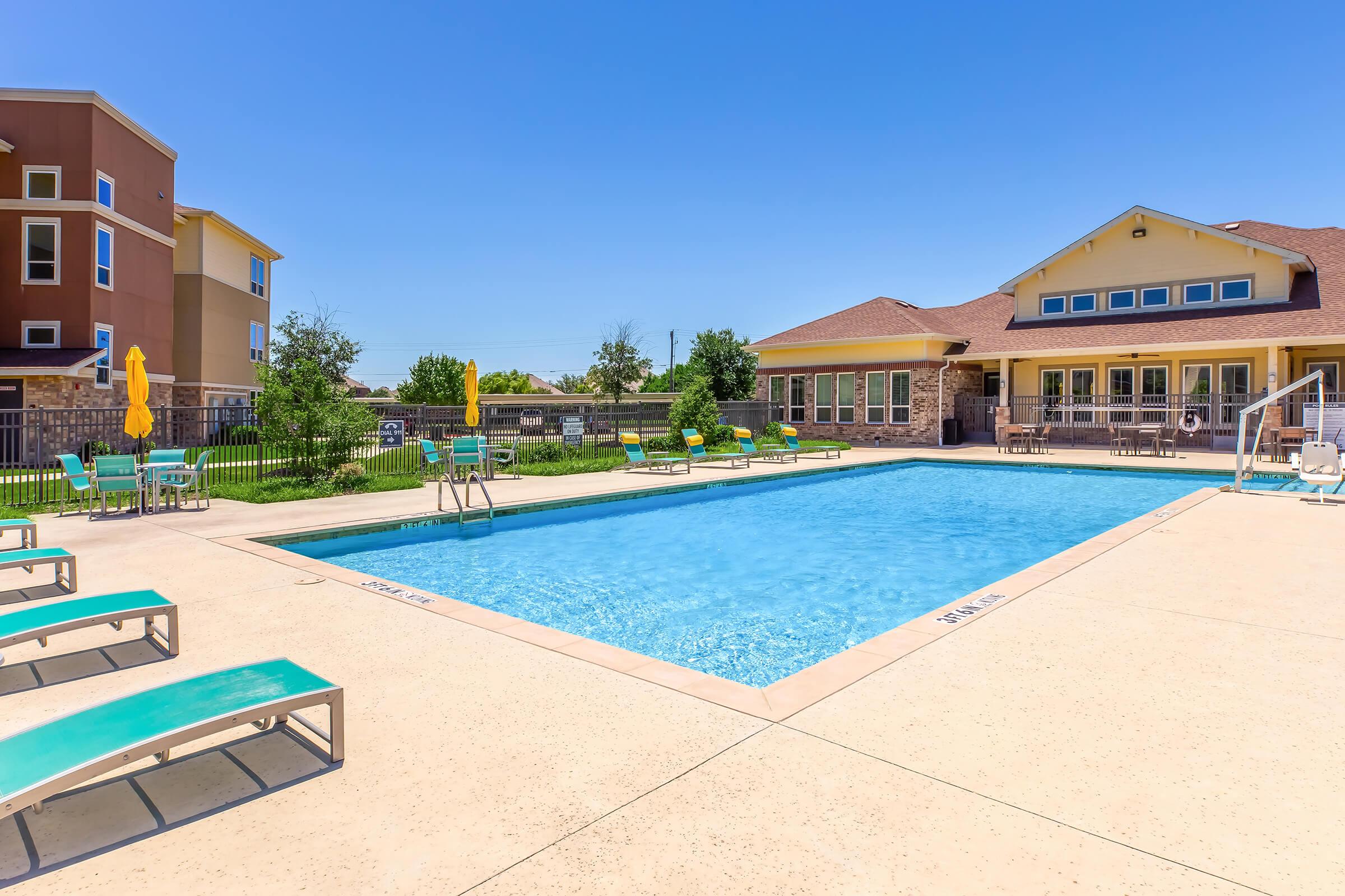 A clear blue swimming pool surrounded by lounge chairs and yellow umbrellas in a sunny outdoor space. In the background, there is a building with a light-colored facade, and a well-maintained lawn with trees and shrubs. The scene conveys a relaxing and inviting atmosphere.