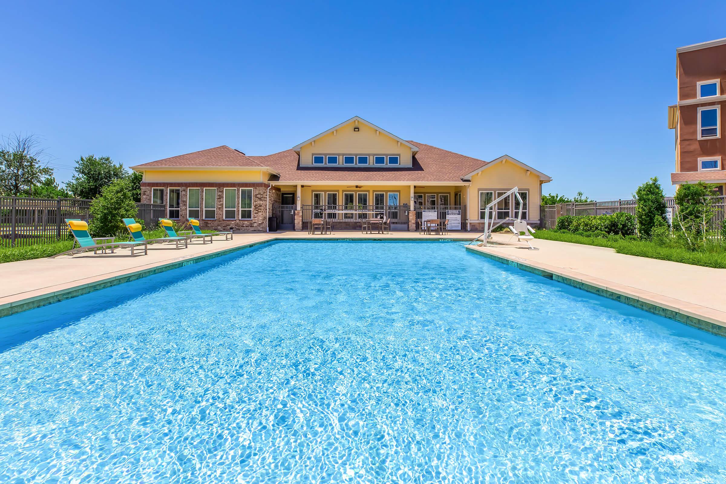 A clear blue swimming pool in the foreground with lounge chairs along the edge. In the background, a large yellow building with a patio area and multiple windows is visible. The sky is bright blue, indicating a sunny day. Lush green grass and trees frame the scene, enhancing the relaxing atmosphere.