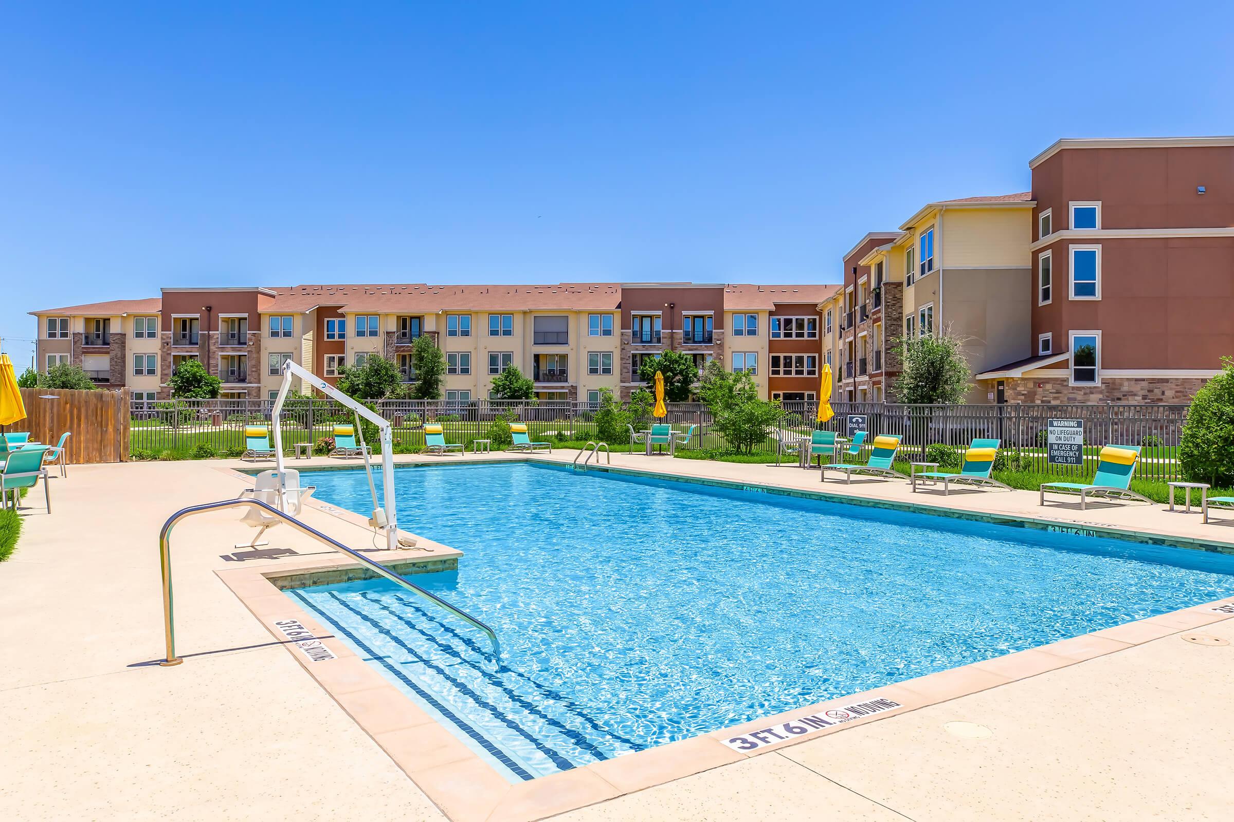 A clear blue swimming pool surrounded by lounge chairs and umbrellas, with an apartment complex in the background. The sky is bright and sunny, creating a vibrant atmosphere. Ideal for relaxation and leisure activities.