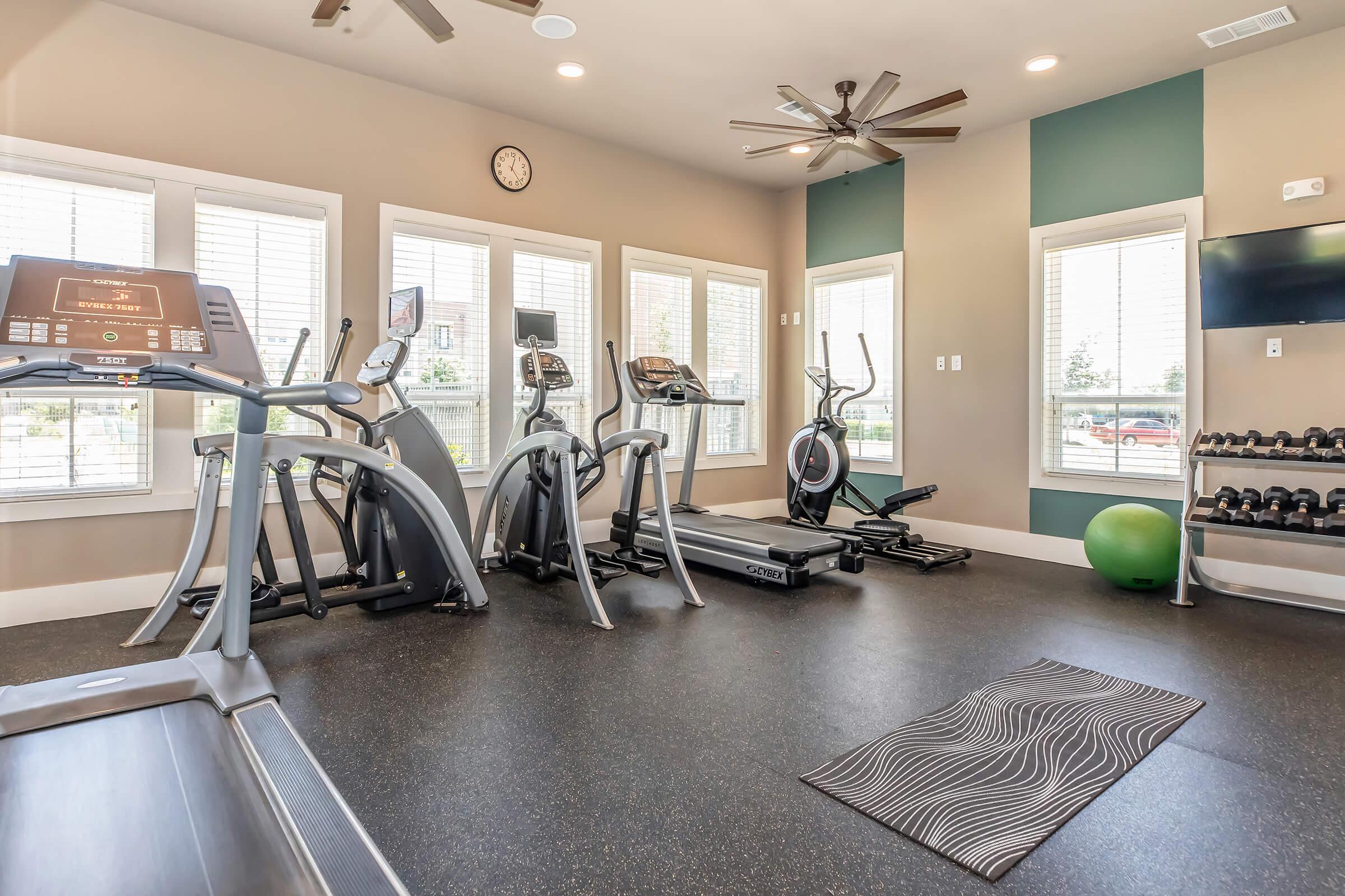 A modern gym interior featuring various exercise equipment, including treadmills, ellipticals, and dumbbells. Large windows provide natural light, and a clock is mounted on the wall. A green exercise ball and yoga mat are also visible on the floor, creating a welcoming fitness space.