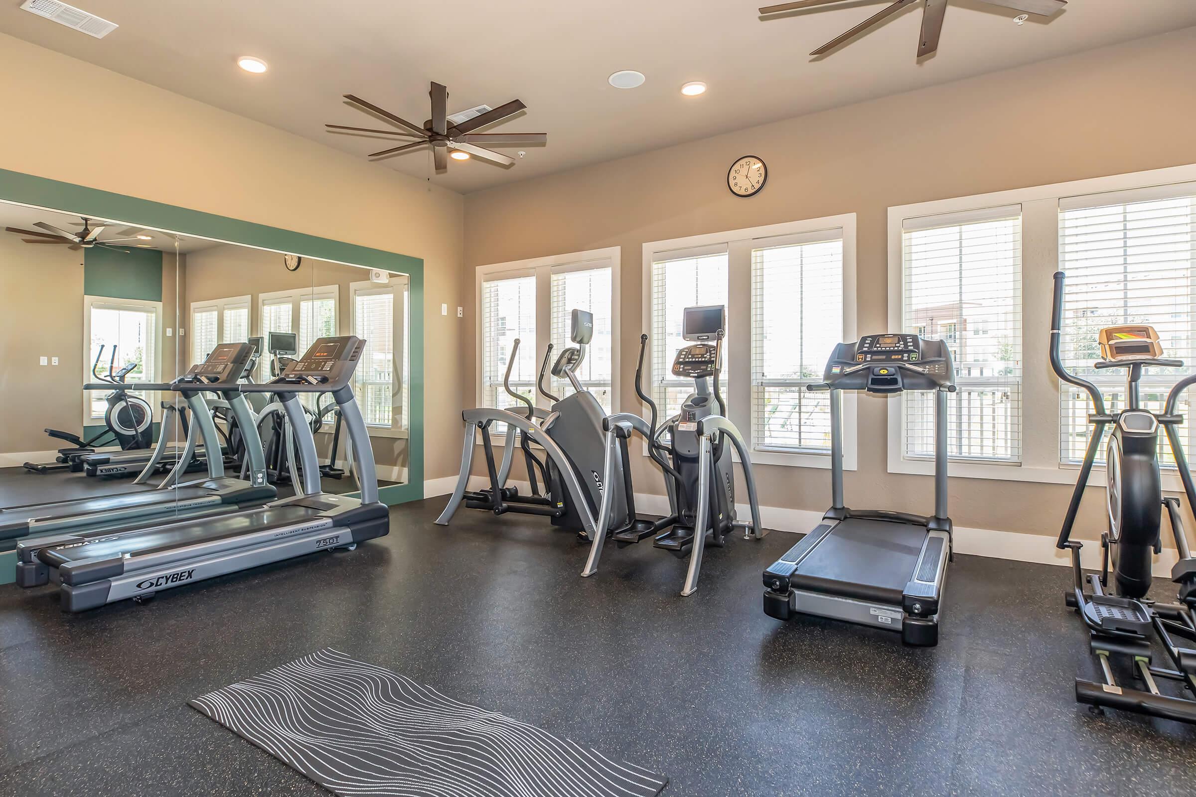 A modern gym area featuring several cardio machines including treadmills and ellipticals, a yoga mat on the floor, large windows with blinds allowing natural light, and ceiling fans. A clock is mounted on the wall, and the space has a clean, minimalistic design.