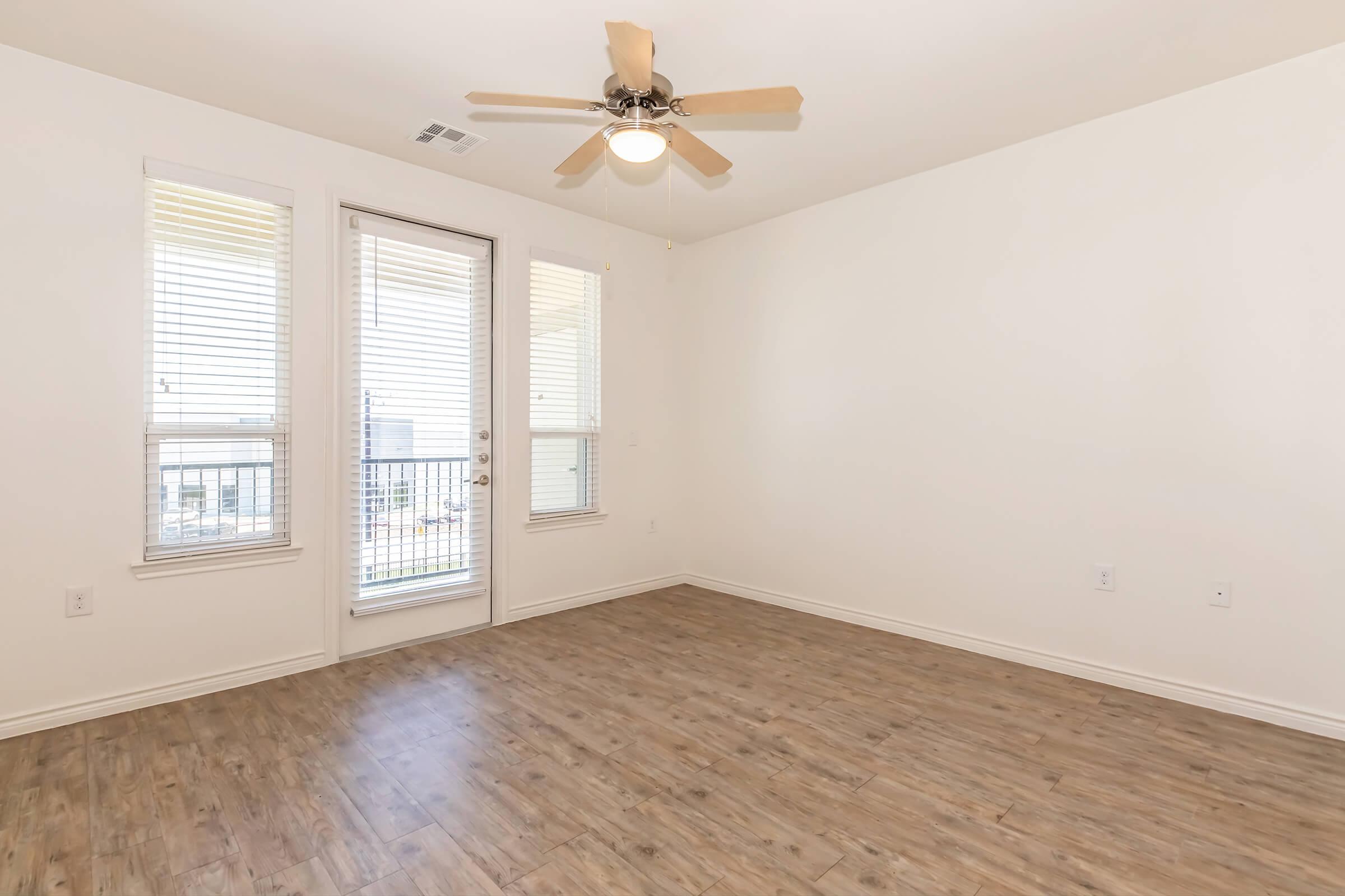 Empty room with light wood flooring, a ceiling fan, and two large windows allowing natural light. A glass door leads to a balcony, and the walls are painted white, creating a bright and airy atmosphere.