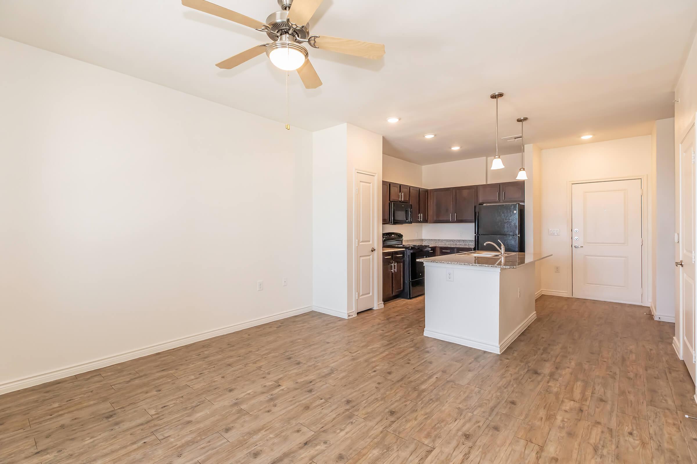 A spacious, modern kitchen and living area featuring light wood-like flooring, a ceiling fan, and dark wooden cabinetry. The kitchen includes black appliances and a breakfast bar. Natural light fills the room, and there’s an entry door visible in the background.