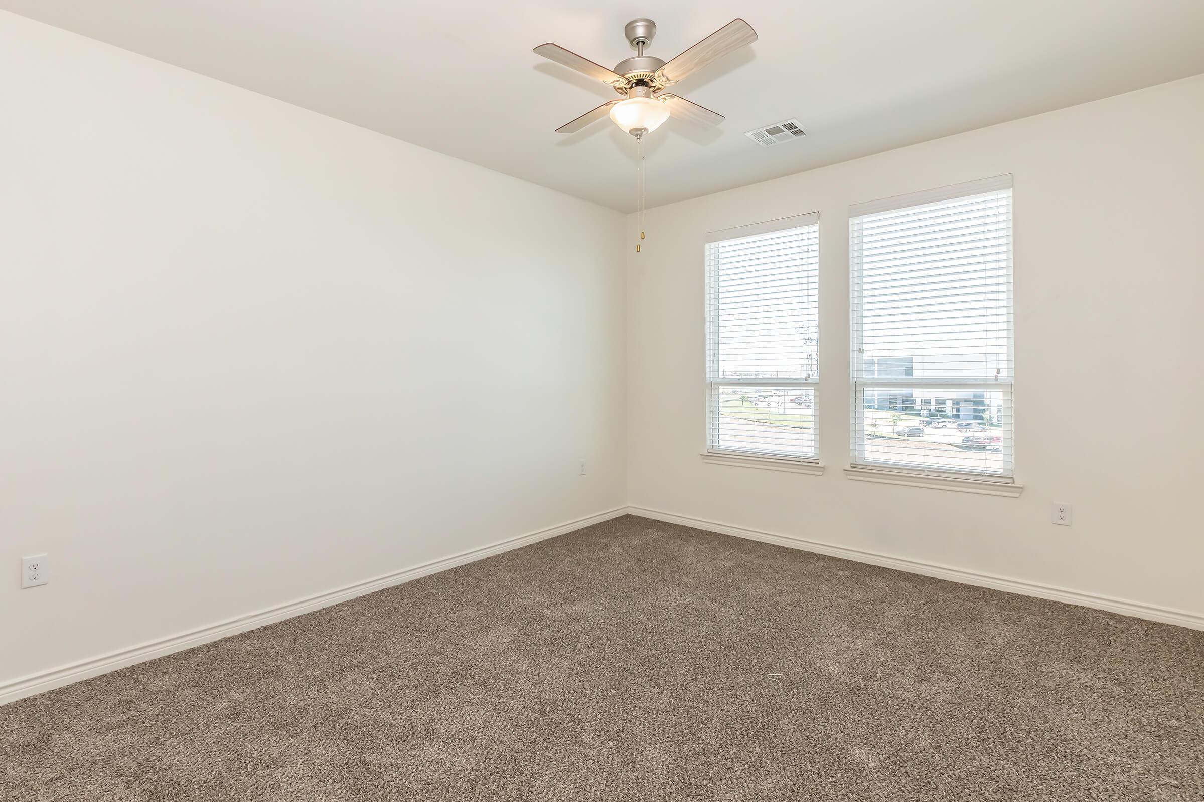 Empty room with beige carpet, white walls, and two windows with blinds allowing natural light. A ceiling fan is mounted in the center, creating a clean and spacious atmosphere.