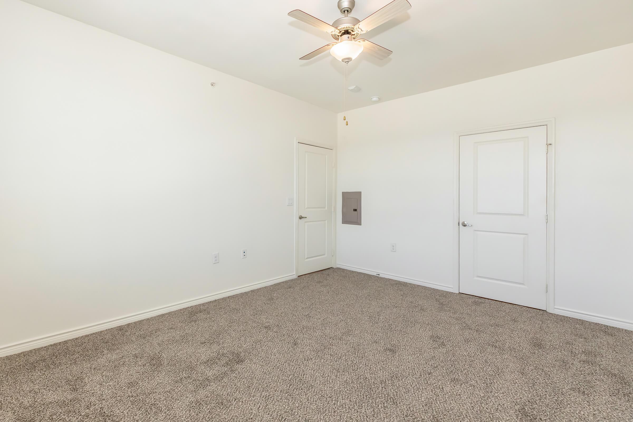 A spacious, empty room with light-colored walls and a plush carpet. A ceiling fan is mounted in the center, and there are two doors on one side, one being a closet. Natural light fills the space, creating a bright and airy atmosphere.